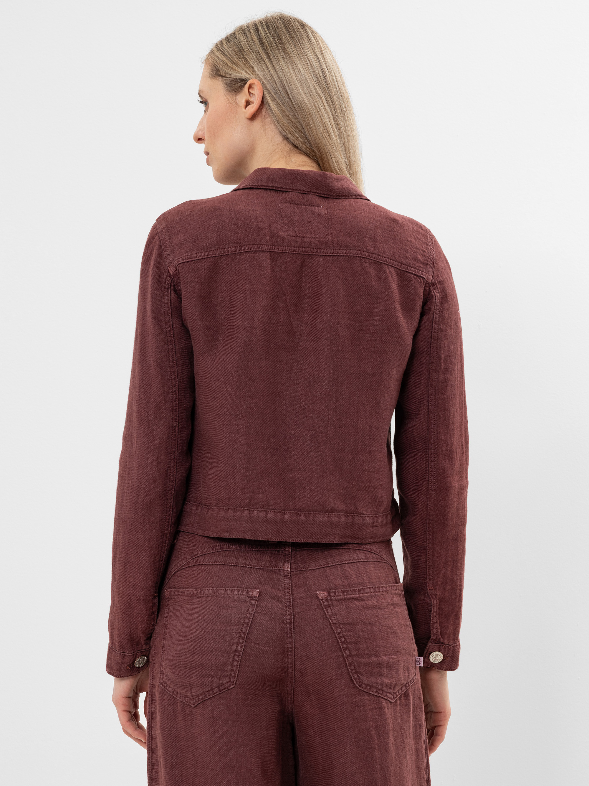 Eine Frau mit glattem, blondem Haar trägt eine kastanienbraune Jeansjacke und eine passende Hose, die von hinten vor einem schlichten weißen Hintergrund gezeigt wird.