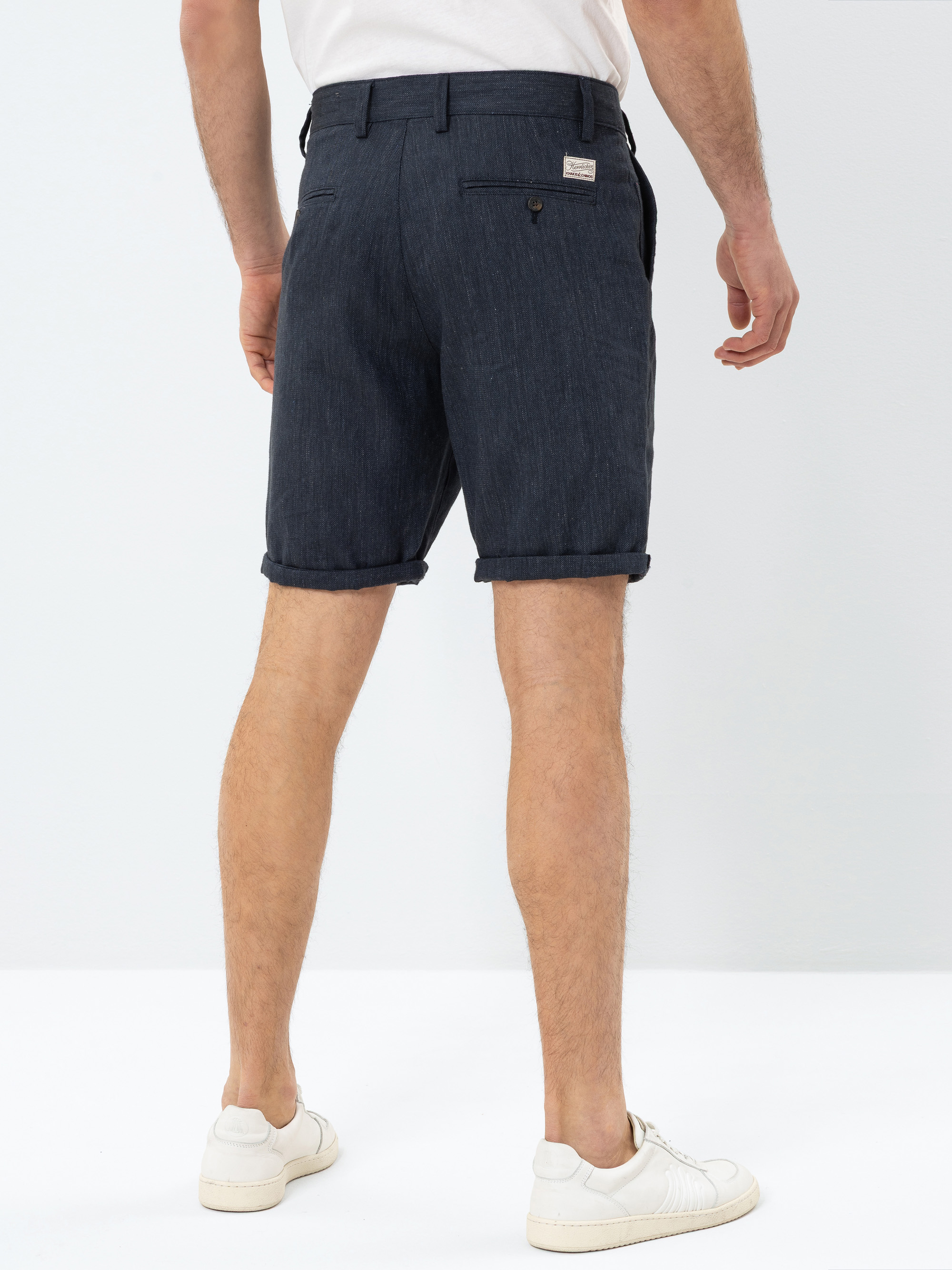 Eine Person steht mit dem Gesicht nach hinten, trägt dunkelblaue Shorts mit gekrempelten Manschetten, ein weißes Hemd und weiße Turnschuhe vor einem schlichten weißen Hintergrund.
