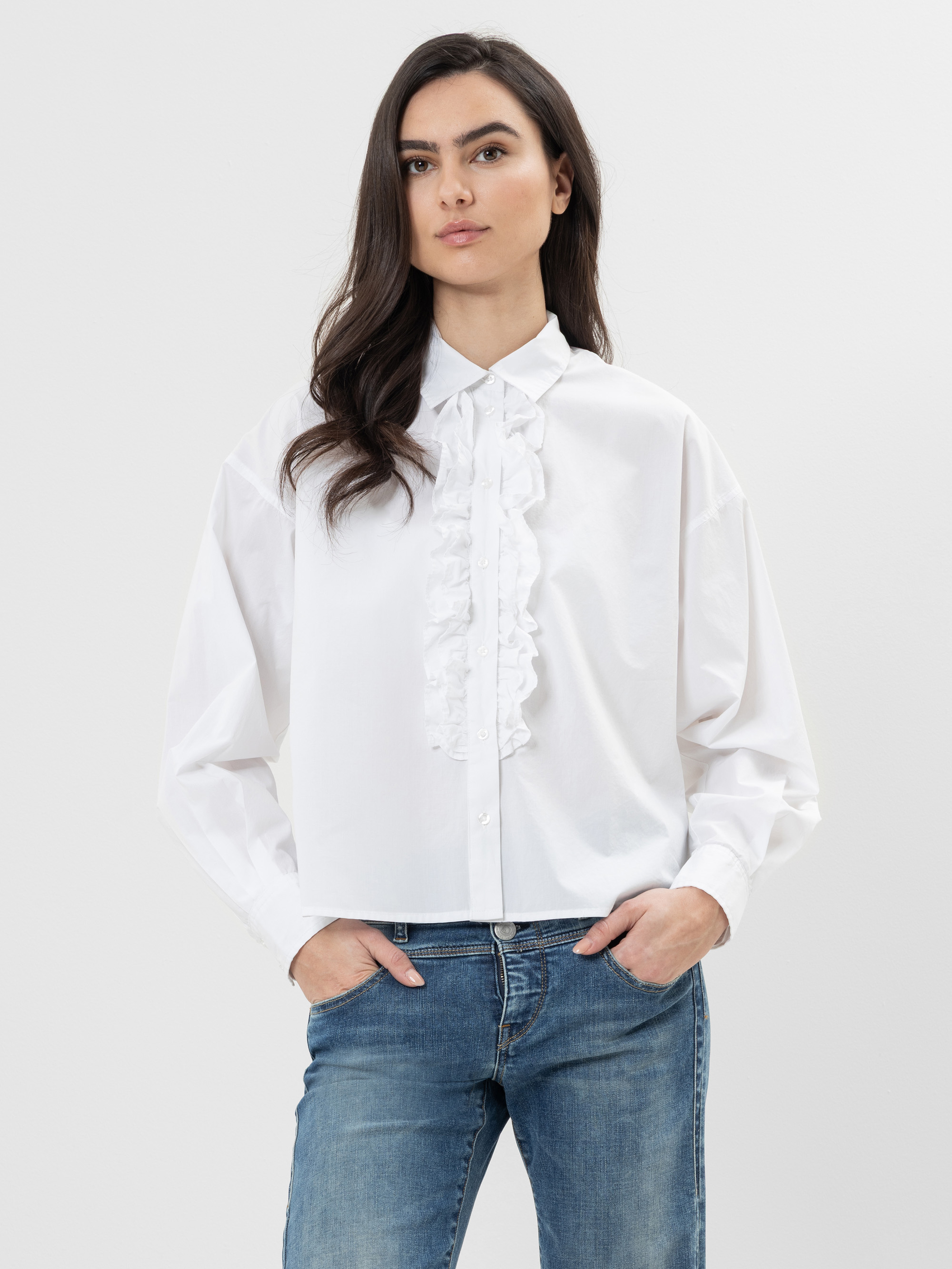 Eine Frau mit langen braunen Haaren trägt eine weiße Button-up-Bluse mit Rüschendetails auf der Vorderseite und blaue Jeans. Sie steht vor einem schlichten hellen Hintergrund, hat die Hände in den Taschen und blickt selbstbewusst in die Kamera.