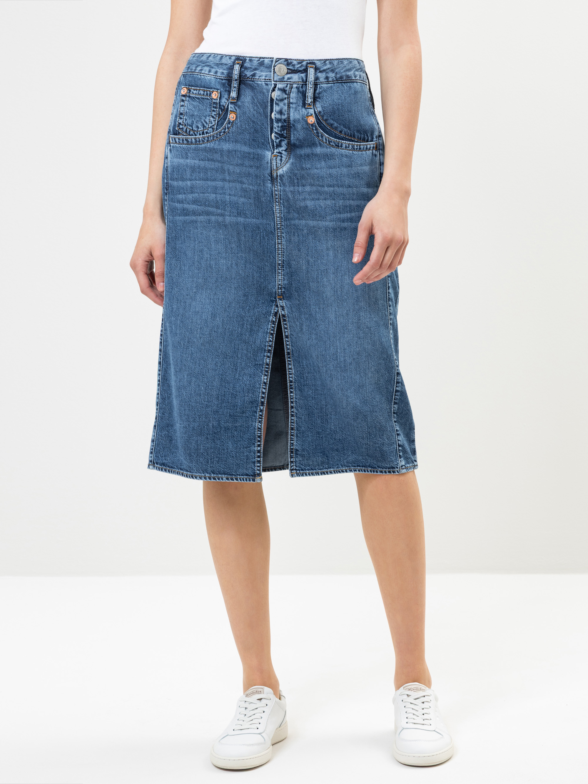 Eine Person, die ein weißes Oberteil, einen knielangen blauen Jeansrock mit einem Schlitz an der Vorderseite und weiße Turnschuhe trägt, steht vor einem schlichten hellen Hintergrund. Das Foto zeigt den Rock von der Taille bis unterhalb der Knie.