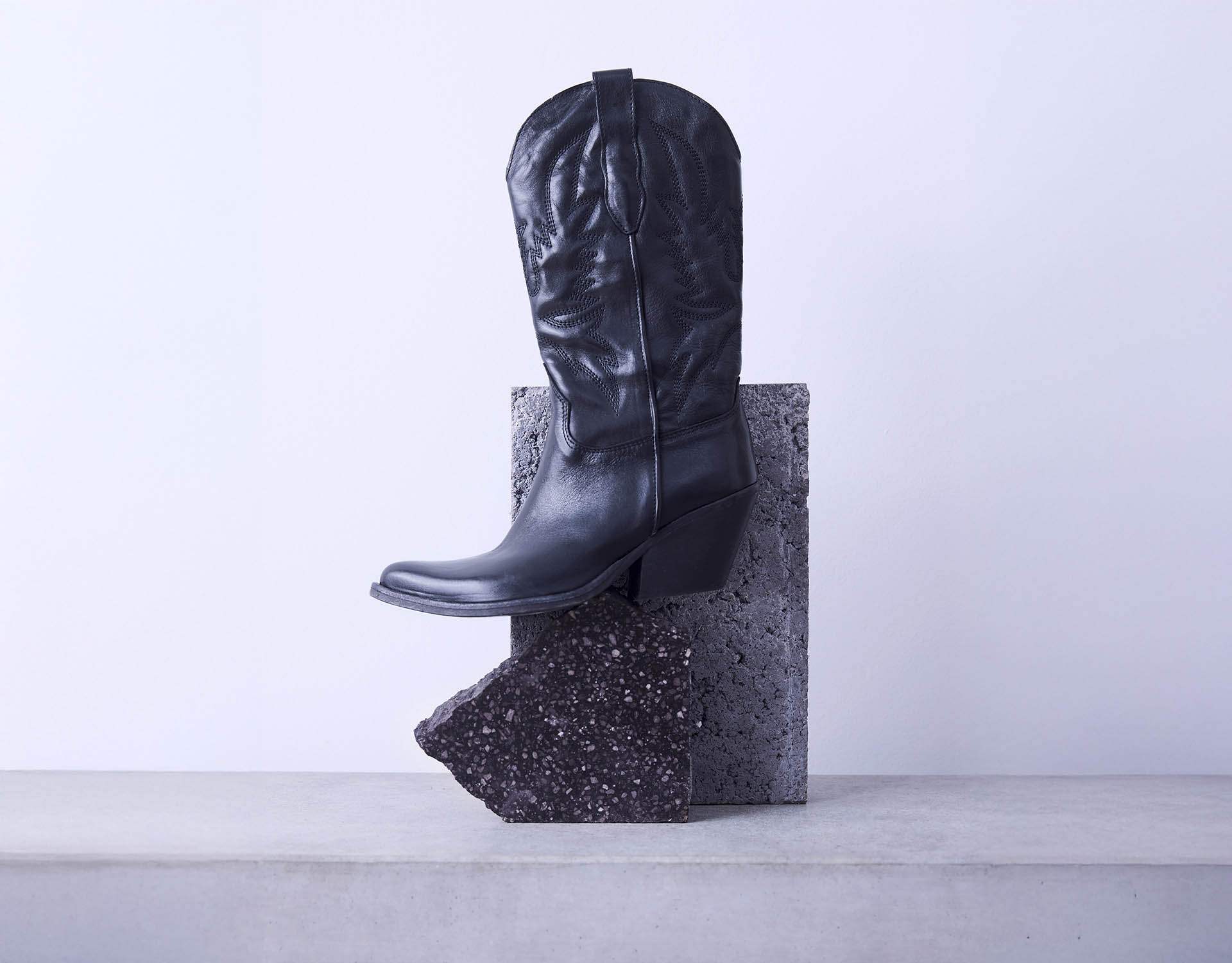 Ein einzelner schwarzer Cowboystiefel steht aufrecht auf einem strukturierten dunklen Stein, hinter dem sich ein rechteckiger grauer Block befindet, vor einem hellgrauen Hintergrund.