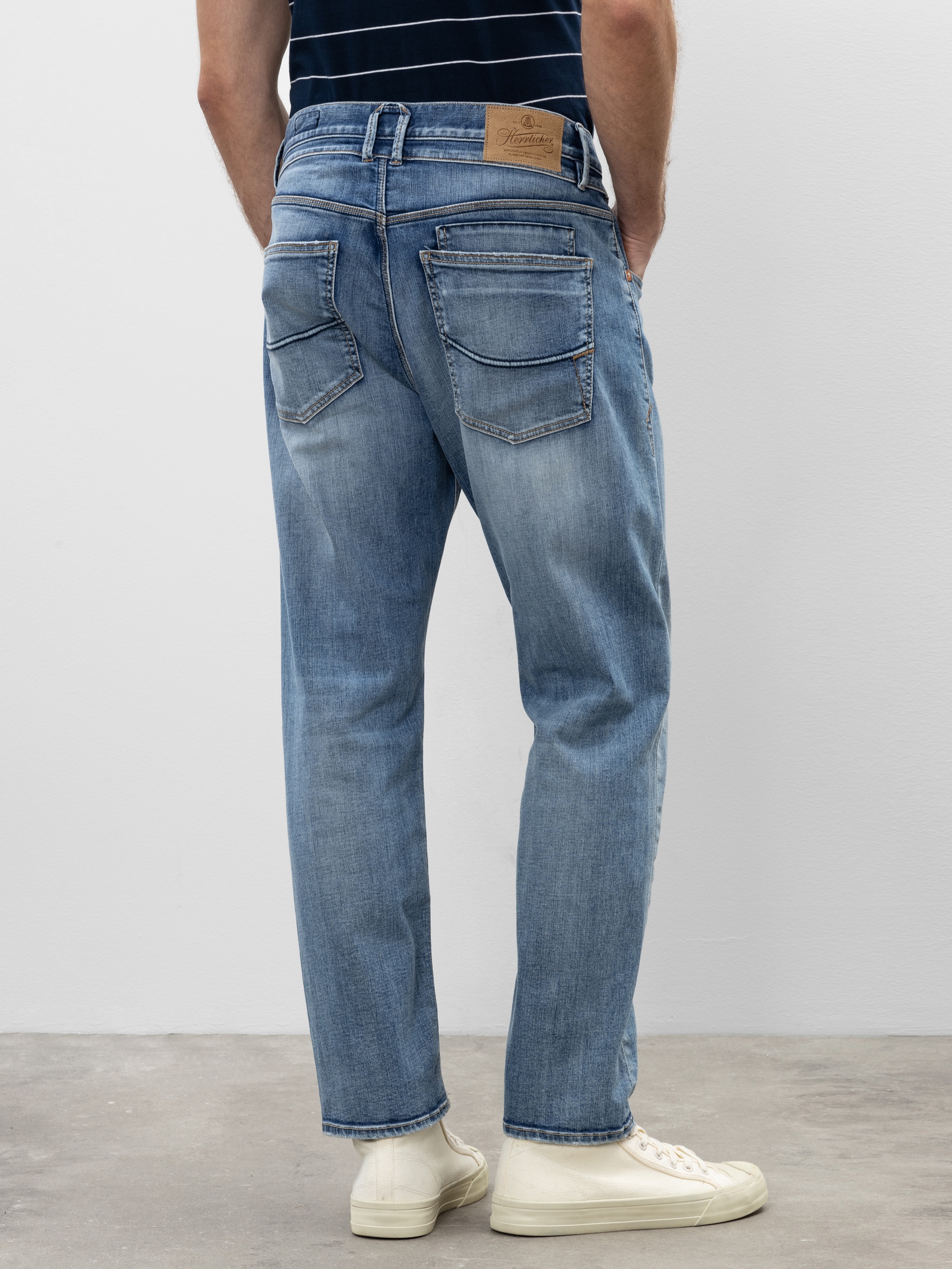 Eine Person, die hellblaue Jeans, ein dunkles Hemd mit weißen Streifen und weiße Turnschuhe trägt, steht auf einem Betonboden vor einer weißen Wand, von der Hüfte abwärts und von hinten gesehen.