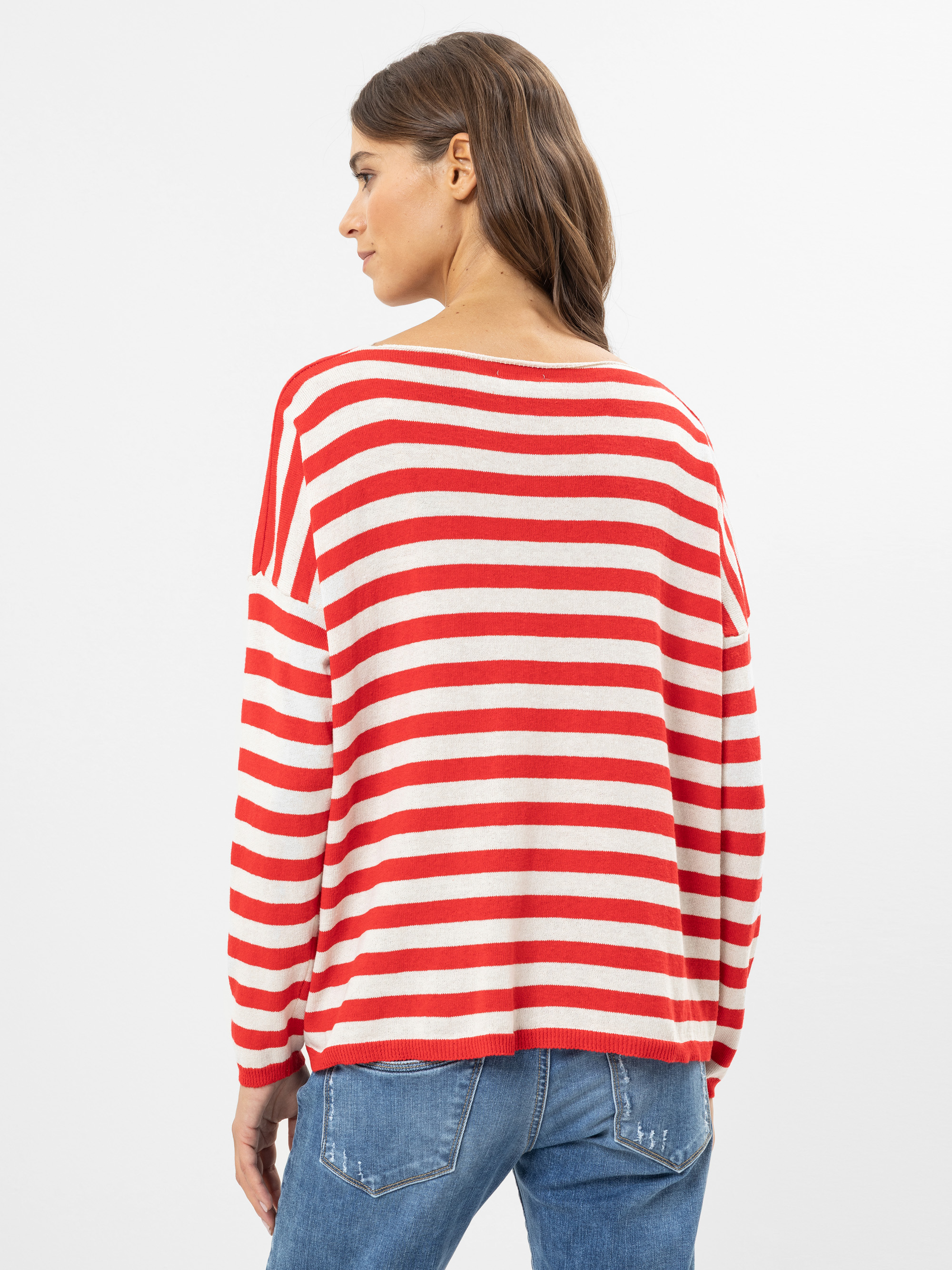Eine Frau steht mit dem Rücken zur Kamera, trägt einen rot-weiß gestreiften langärmeligen Pullover und blaue Jeans vor einem schlichten weißen Hintergrund.