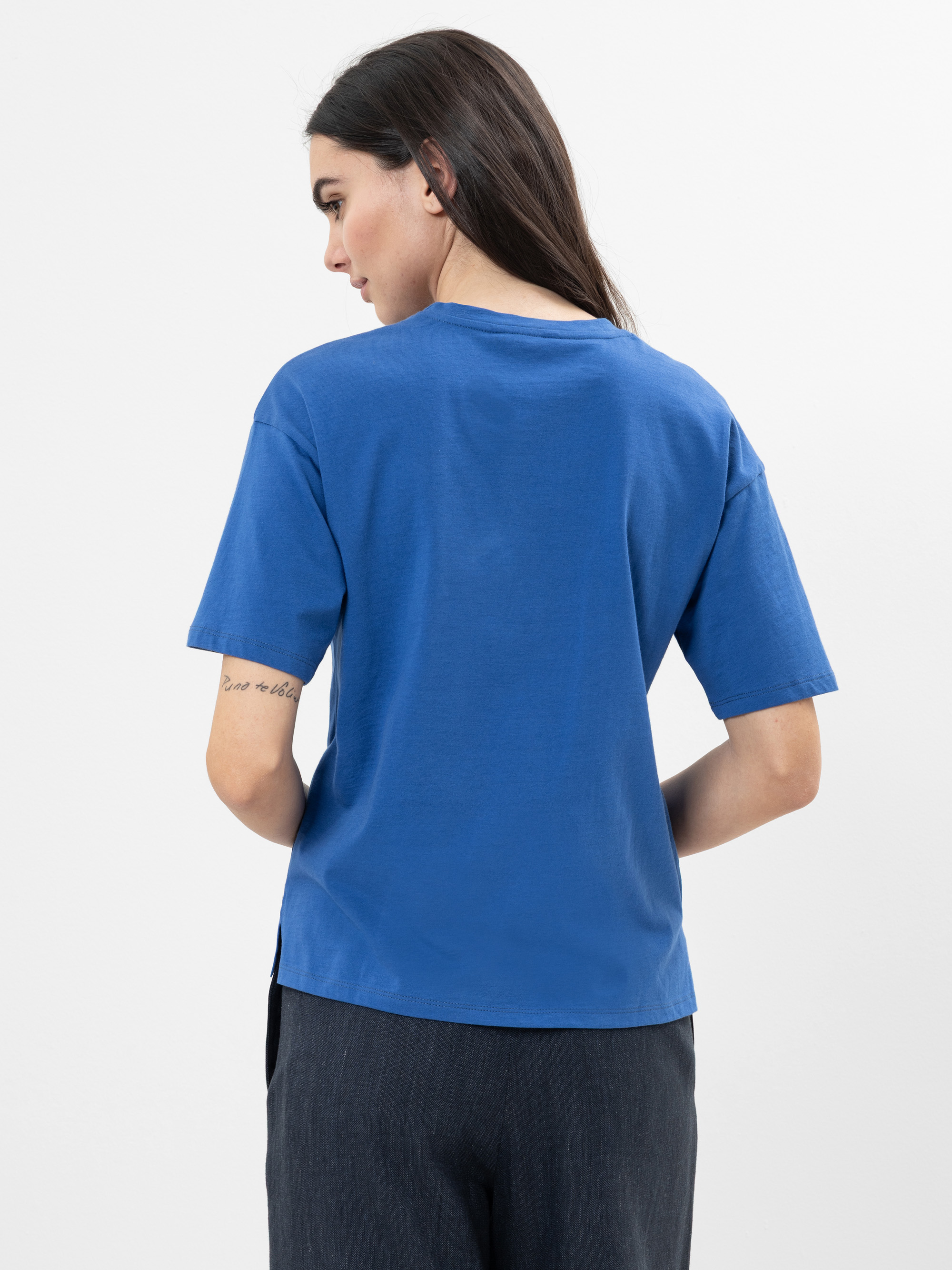 Frau mit langen dunklen Haaren, abgewandt stehend, bekleidet mit einem blauen kurzärmeligen T-Shirt und einer dunklen Hose, die eine Tätowierung auf ihrem linken Unterarm zeigt, vor einem schlichten weißen Hintergrund.