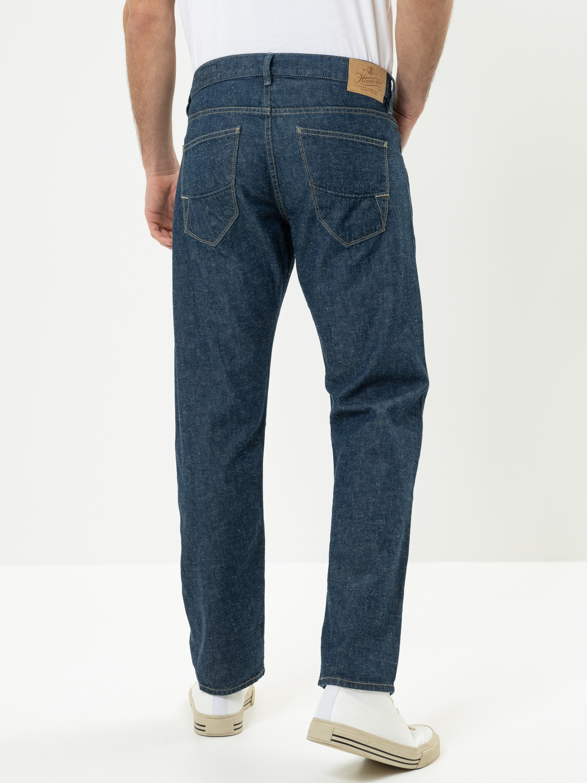 Eine Person, die blaue Jeans und weiße Turnschuhe trägt, steht mit dem Rücken zur Kamera vor einem schlichten weißen Hintergrund. Die Jeans hat zwei Gesäßtaschen und einen Lederaufnäher am Bund.
