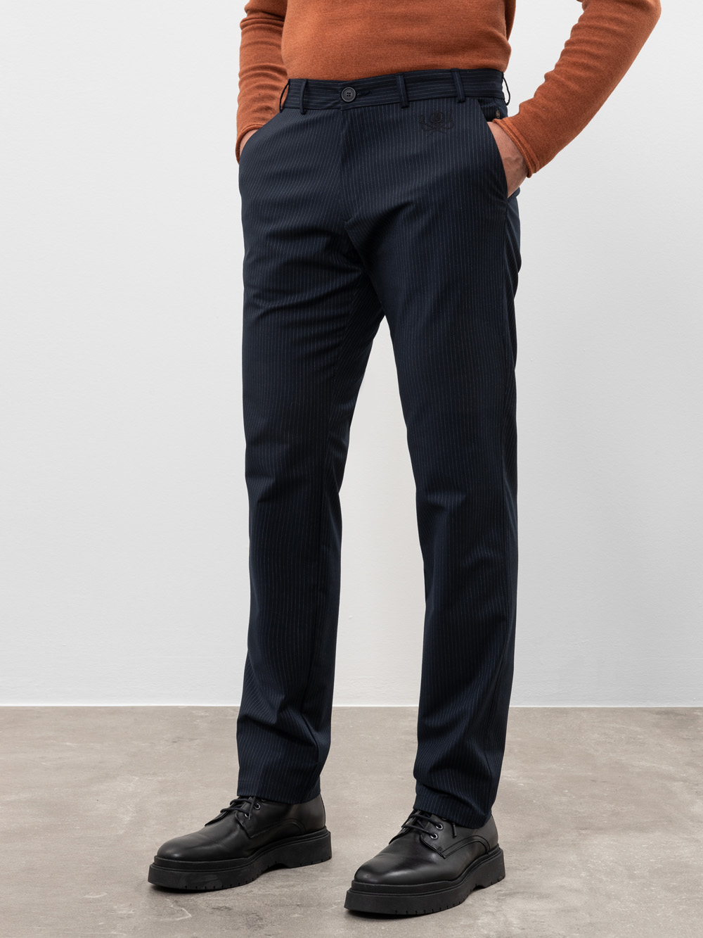 Eine Person, die ein rostfarbenes langärmeliges Hemd, eine dunkle Nadelstreifenhose und schwarze Anzugschuhe trägt, steht mit den Händen in den Taschen vor einer hellen Wand. Ihr Kopf ist nicht zu sehen.
