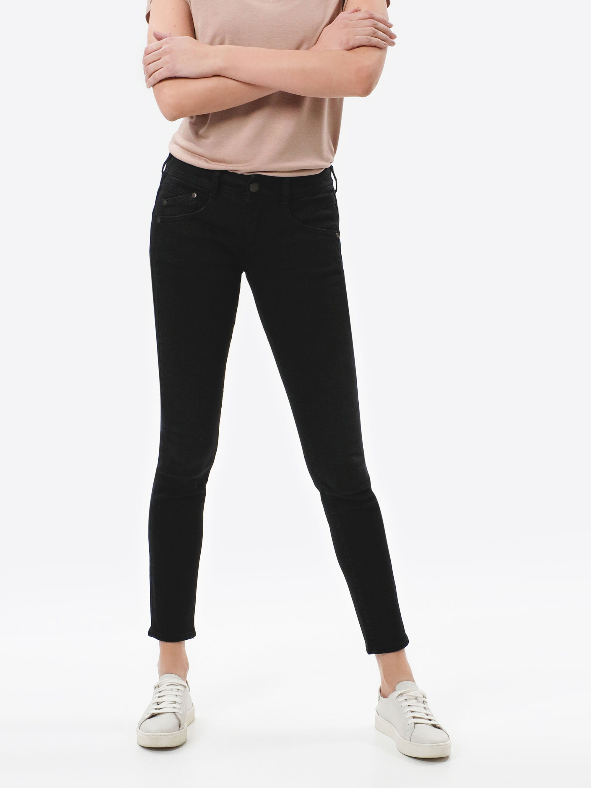 Eine Person steht mit verschränkten Armen, einem hellrosa T-Shirt, schwarzen Skinny-Jeans und weißen Turnschuhen vor einem schlichten weißen Hintergrund. Ihr Gesicht ist nicht zu sehen.