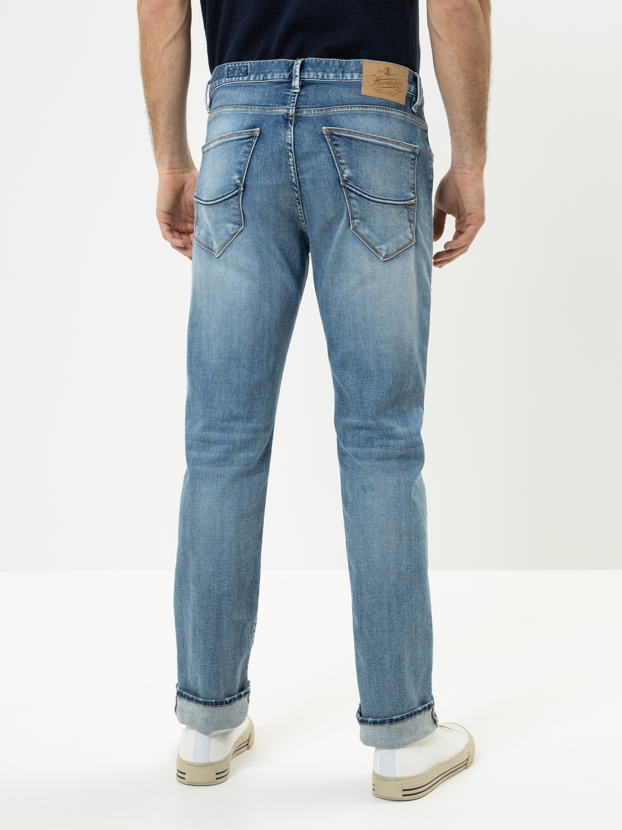 Eine Person steht mit dem Gesicht nach unten und trägt eine hellblaue Jeans mit aufgerollten Bündchen und weiße Turnschuhe. Die Jeans hat zwei Gesäßtaschen und einen hellbraunen Aufnäher am Bund. Der Hintergrund ist einfarbig und weiß.