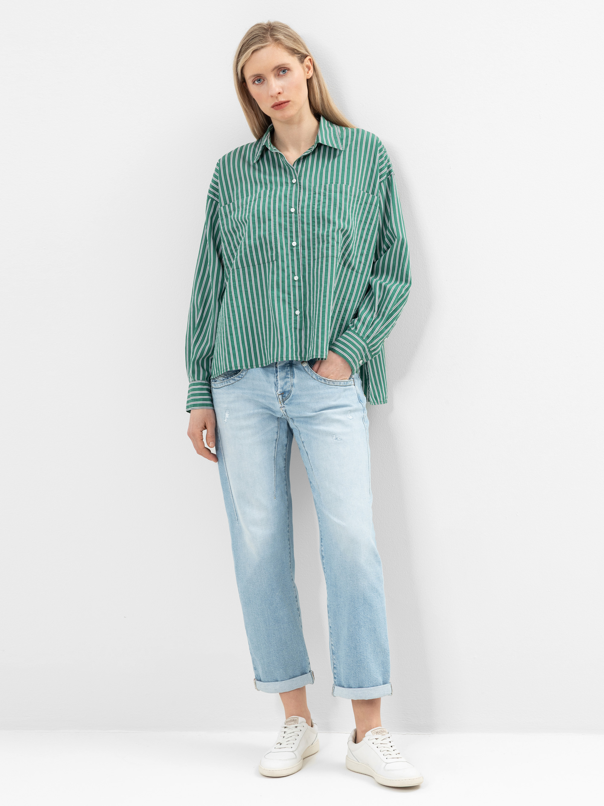 Eine Frau steht vor einer schlichten weißen Wand. Sie trägt ein grün-weiß gestreiftes Button-up-Hemd, hellblaue Jeans und weiße Turnschuhe. Sie hat glattes blondes Haar und einen neutralen Gesichtsausdruck.