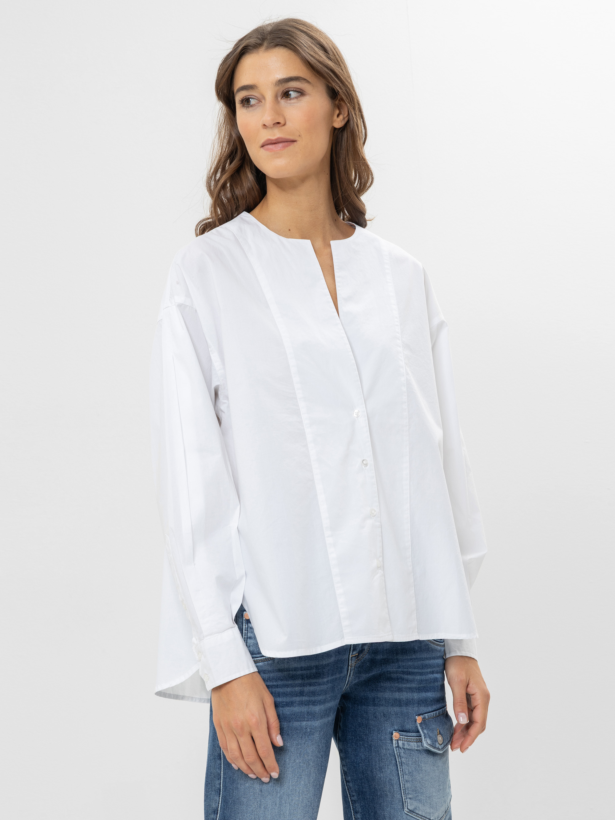 Eine Frau mit langen braunen Haaren, die eine locker sitzende weiße Button-up-Bluse und blaue Jeans trägt, steht vor einem schlichten weißen Hintergrund, schaut leicht zur Seite und lächelt sanft.