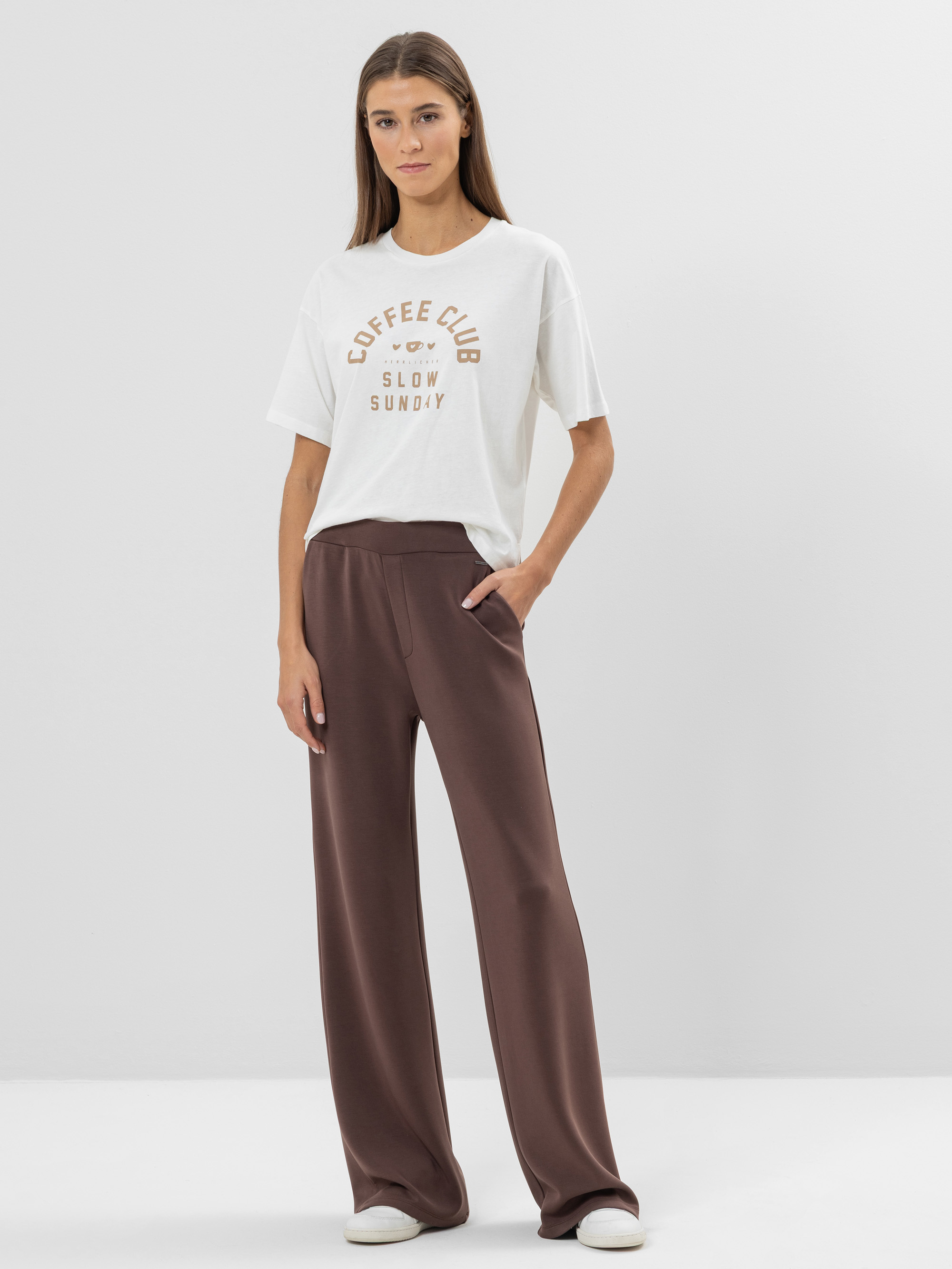 Eine Frau steht vor einem schlichten Hintergrund, trägt ein weißes Coffee Club Slow Sunday-T-Shirt, eine braune Hose mit weitem Bein und weiße Turnschuhe, eine Hand in der Tasche und einen entspannten Gesichtsausdruck.