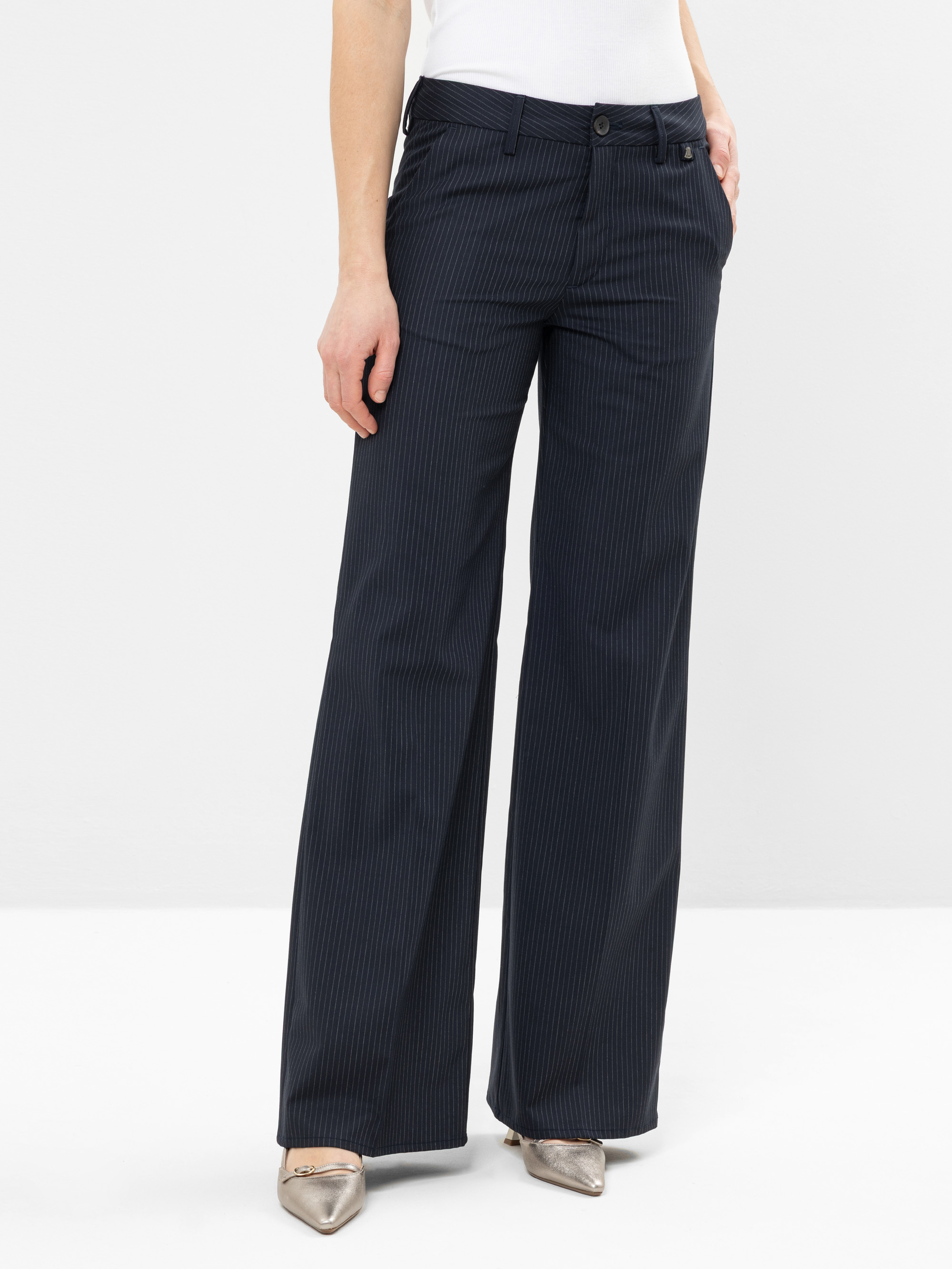 Eine Person, die eine marineblaue, gestreifte Hose mit weitem Bein und Metallic-Flachschuhe mit spitzen Zehen trägt, die Hände in den Taschen, steht vor einem schlichten weißen Hintergrund.