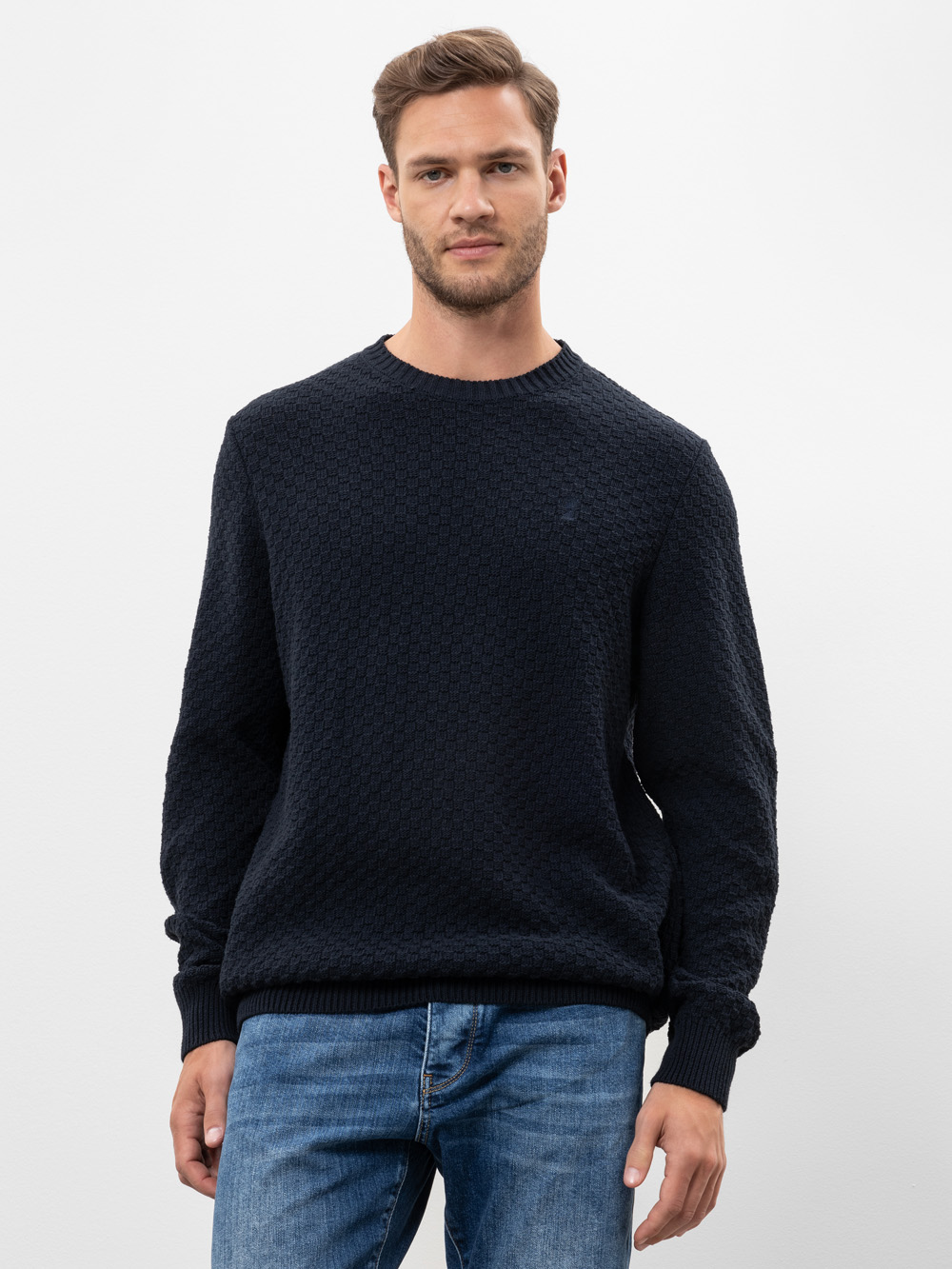 Ein Mann mit kurzen braunen Haaren und gestutztem Bart trägt einen strukturierten dunkelnavyfarbenen Pullover und blaue Jeans und steht vor einem schlichten weißen Hintergrund. Er blickt direkt in die Kamera und hat einen neutralen Blick.