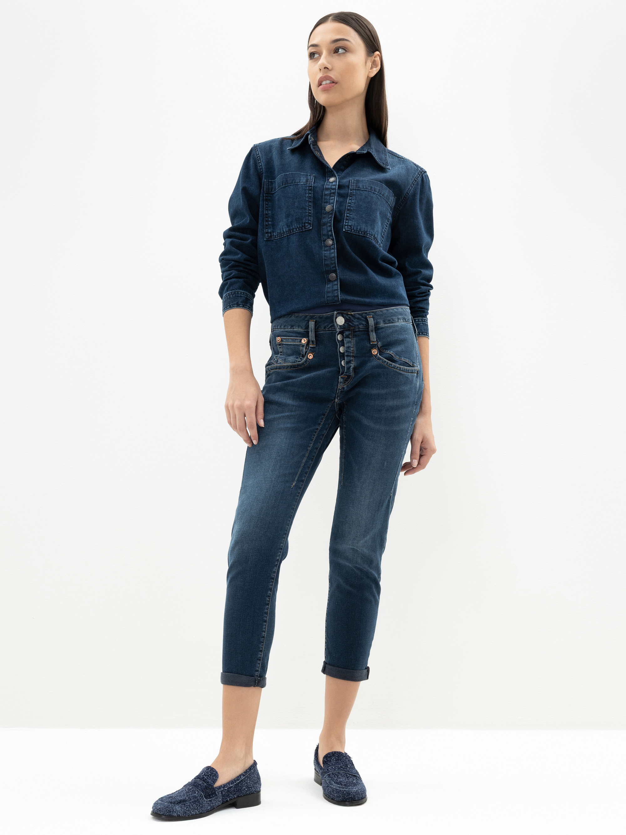 Eine Frau steht vor einem schlichten weißen Hintergrund und trägt ein dunkles Jeanshemd mit Knopfleiste, dazu passende Jeans mit gekrempelten Manschetten und blaue strukturierte Halbschuhe. Sie blickt leicht zur Seite und hat eine Hand in die Hüfte ges