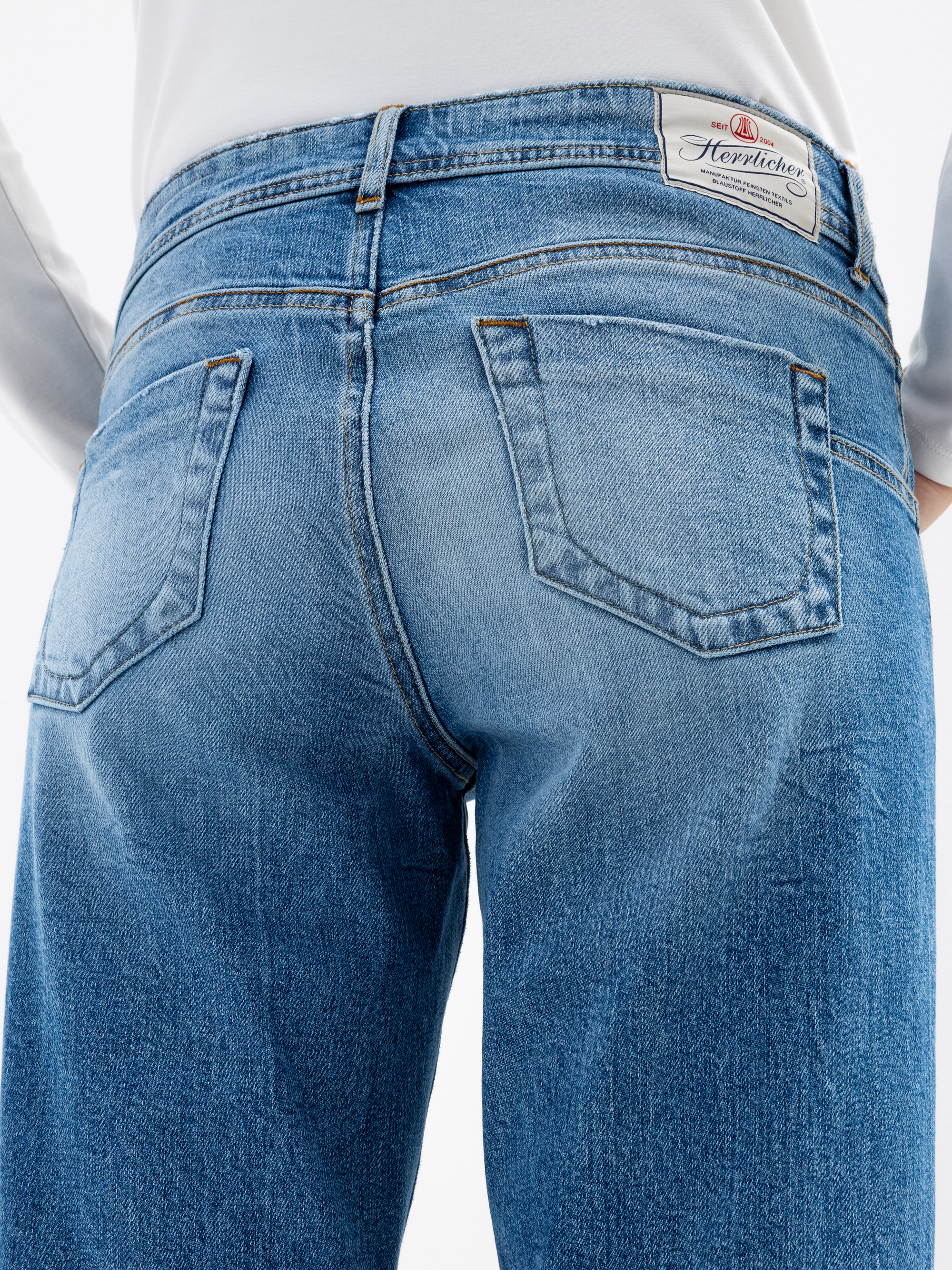 Nahaufnahme des Rückens einer Person, die eine hellblaue Jeans mit zwei Gesäßtaschen und einem weißen Etikett mit der Aufschrift "Herrlicher" am Bund trägt. Die Person trägt außerdem ein weißes Oberteil.