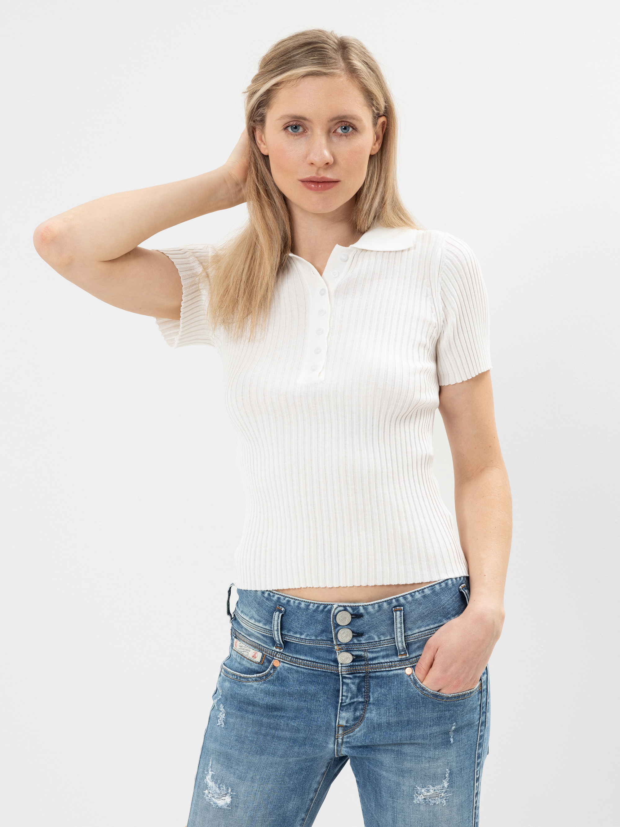 Eine Frau mit blondem Haar, die ein kurzärmeliges, weißes, geripptes Poloshirt und blaue, abgenutzte Jeans trägt, posiert mit einer Hand in ihrem Haar und der anderen in ihrer Tasche vor einem schlichten weißen Hintergrund.