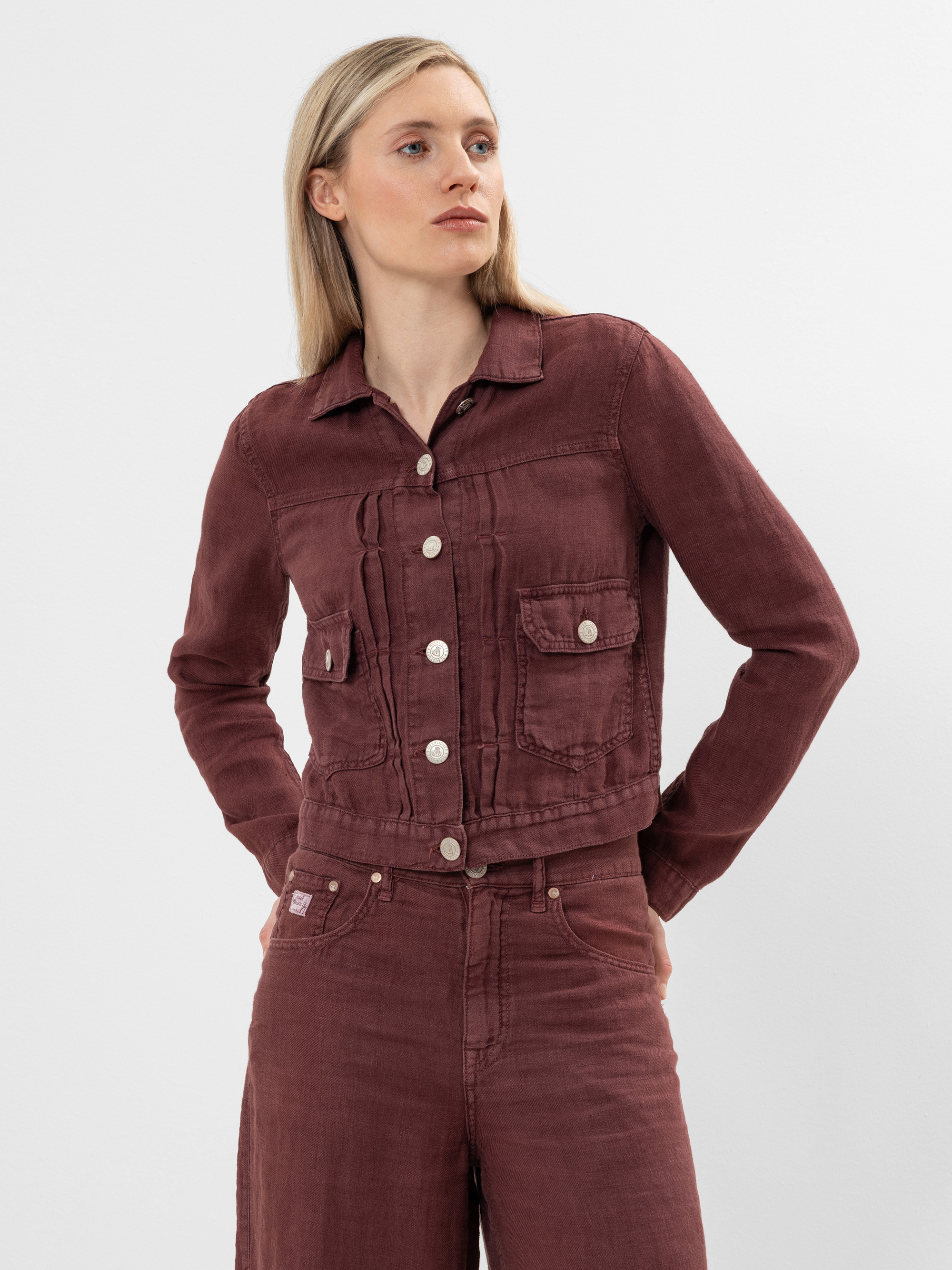 Eine Frau mit blondem Haar steht vor einem unifarbenen Hintergrund und trägt eine passende bordeauxfarbene Jeansjacke und eine Hose mit hoher Taille. Die Jacke hat silberne Knöpfe und Vordertaschen. Sie blickt zur Seite und hat die Hände hinter dem Rü