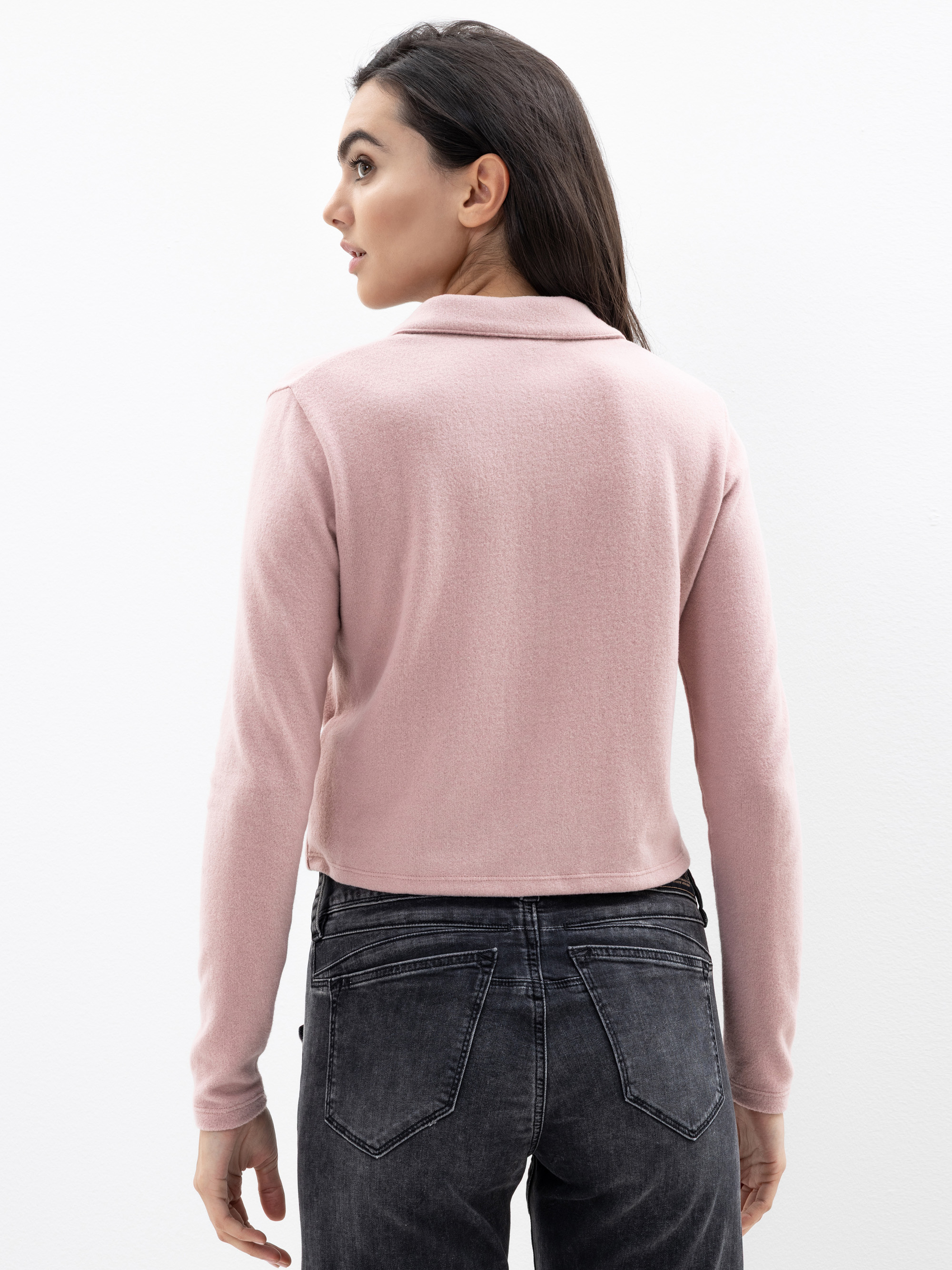 Eine Frau mit langen dunklen Haaren trägt ein hellrosafarbenes, langärmeliges Oberteil und schwarze Jeans. Sie steht vor einem schlichten weißen Hintergrund und wendet ihren Kopf leicht nach links.