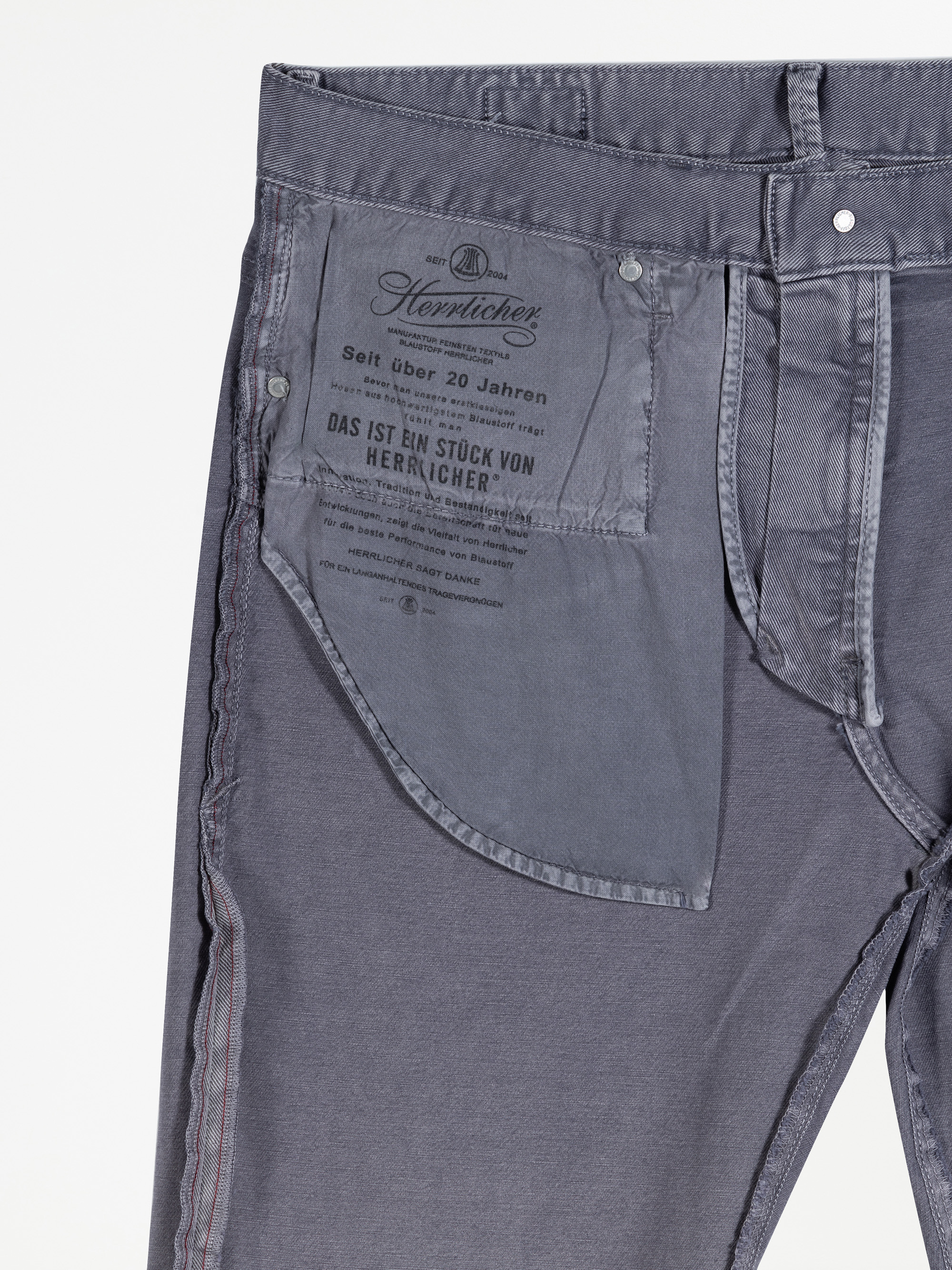 Nahaufnahme einer umgedrehten grauen Hose, die die Innentasche mit aufgedrucktem Branding und Text in deutscher Sprache des Herstellers Herrlicher zeigt. Der Hosenknopf, der Bund und die Nahtdetails sind sichtbar.