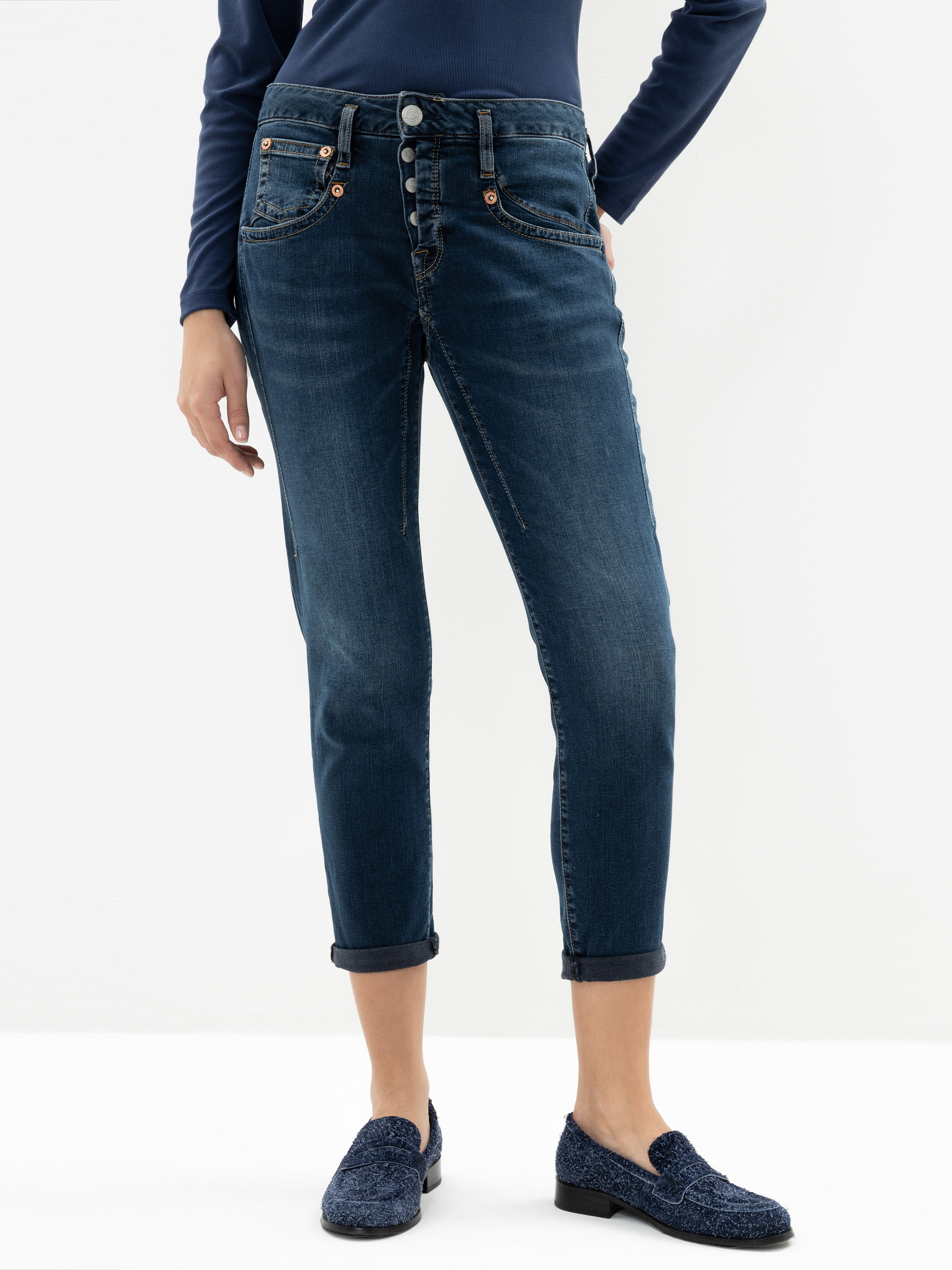 Eine Person, die dunkelblaue Jeans mit Manschetten und sichtbaren Knöpfen, ein marineblaues langärmeliges Oberteil und blaue strukturierte Halbschuhe trägt, steht vor einem schlichten weißen Hintergrund.