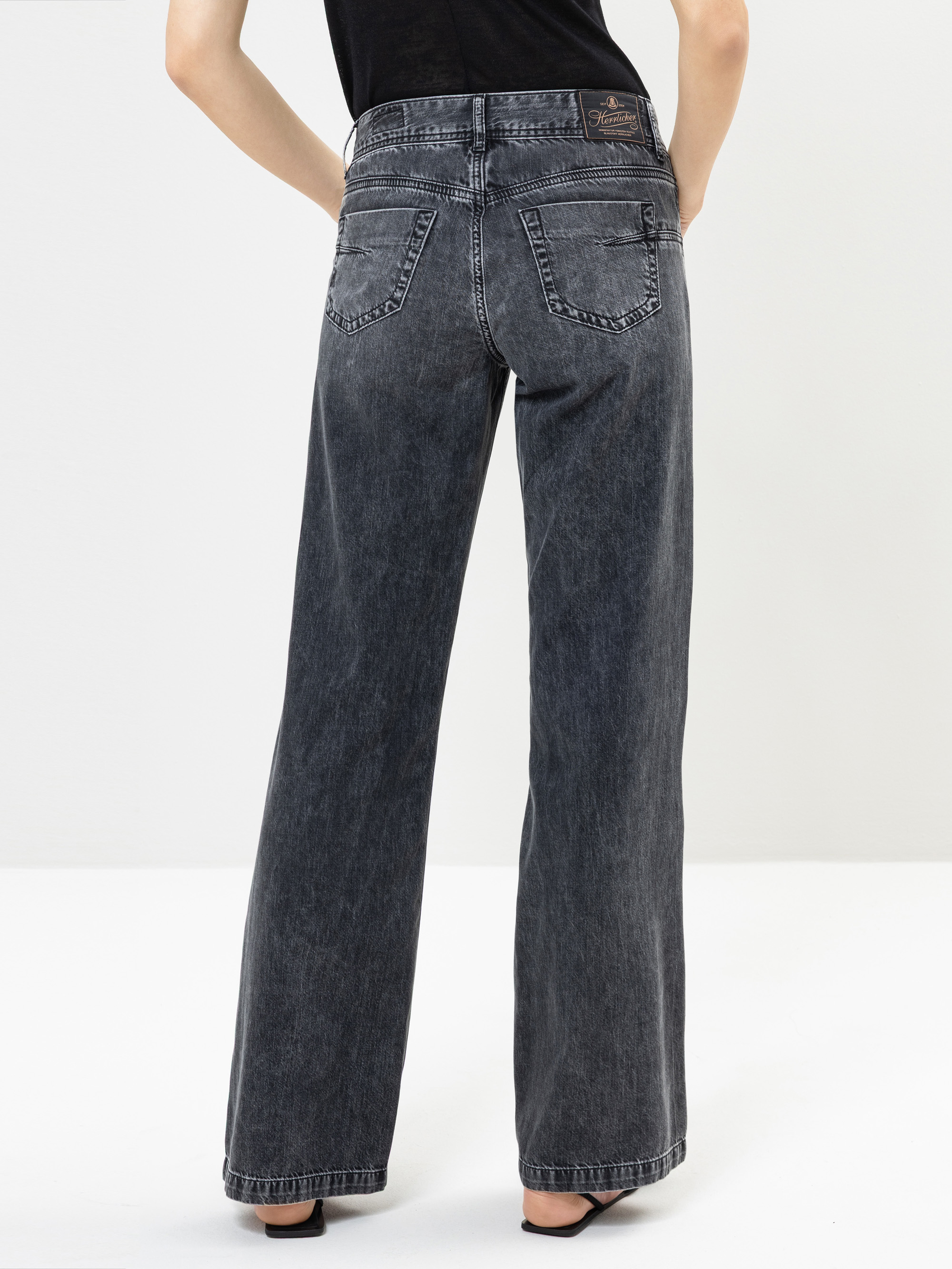 Eine Person, die eine schwarze Jeans mit hoher Taille und weitem Bein sowie schwarze offene Sandalen trägt, steht mit dem Rücken zur Kamera vor einem schlichten weißen Hintergrund.