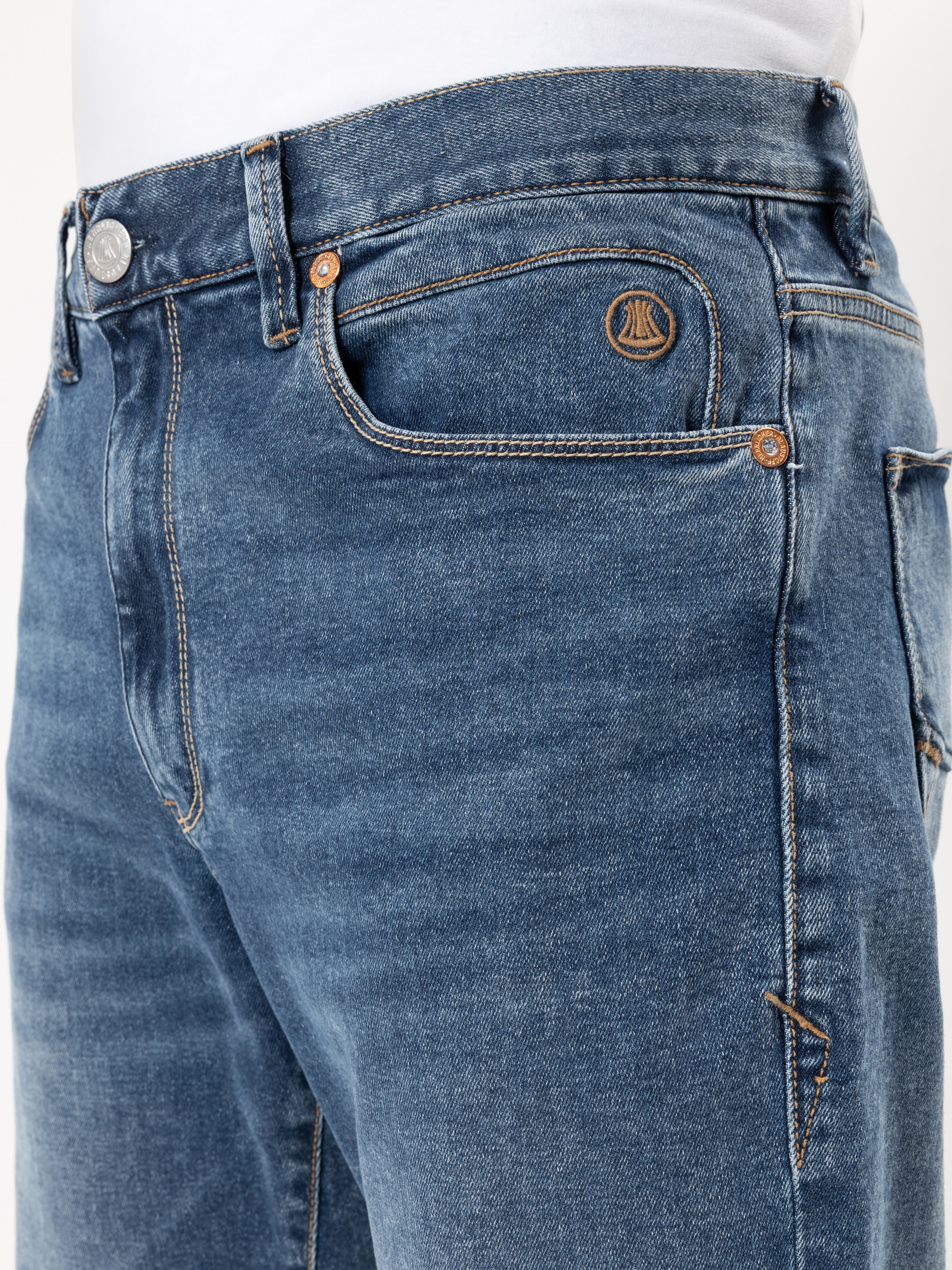 Nahaufnahme einer Person, die eine blaue Denim-Jeans mit hellbraunen Nähten und ein weißes Hemd in die Hose steckt. Die Jeans hat ein kleines gesticktes Logo über der rechten Vordertasche und sichtbare Kupfernieten.