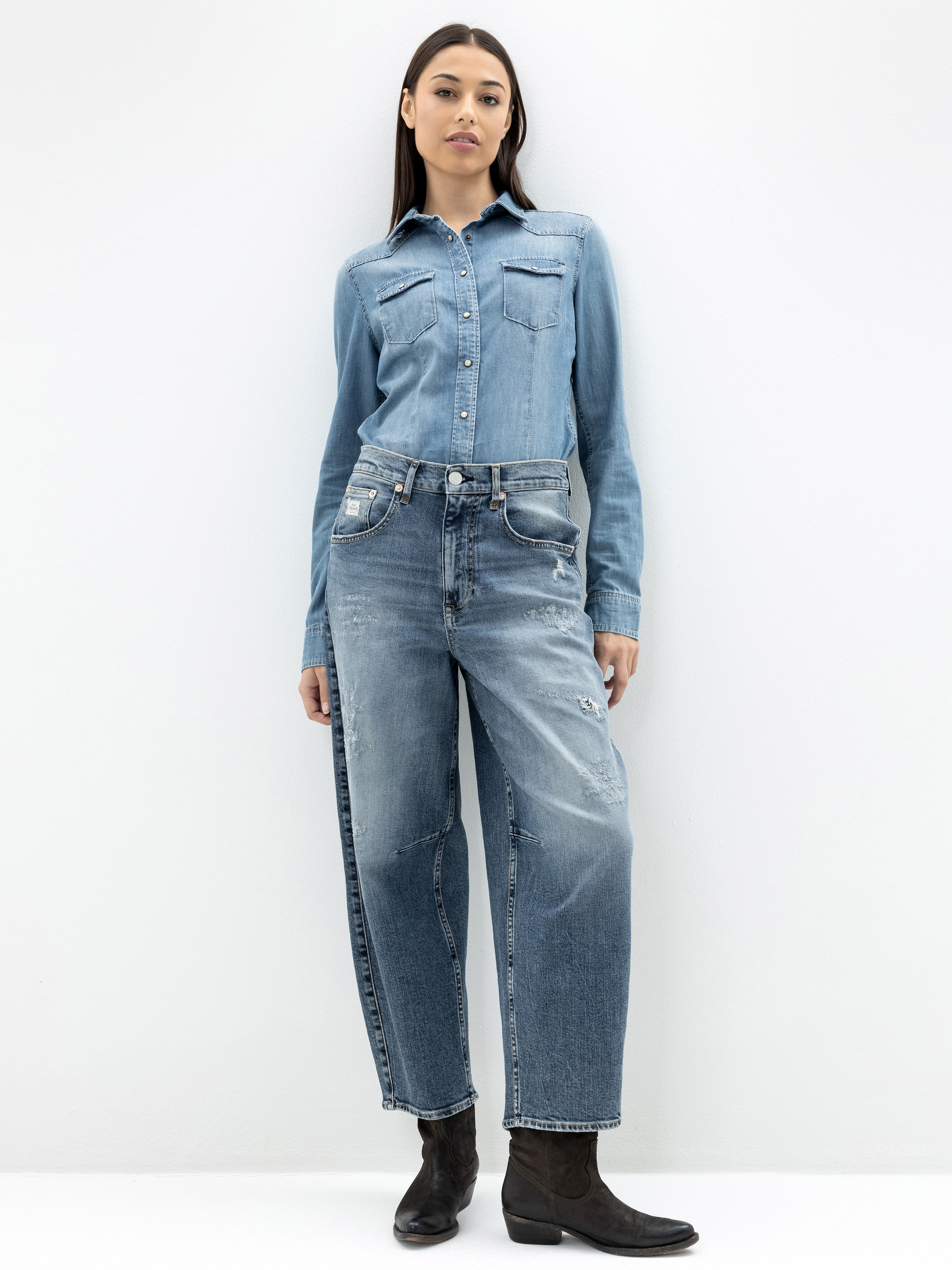 Eine Frau steht vor einer schlichten weißen Wand. Sie trägt ein helles Jeanshemd mit Knopfleiste, das in eine hochtaillierte, locker sitzende Bluejeans mit leichten Gebrauchsspuren gesteckt wird, und dunkle Stiefeletten. Sie schaut mit neutralem Blick d