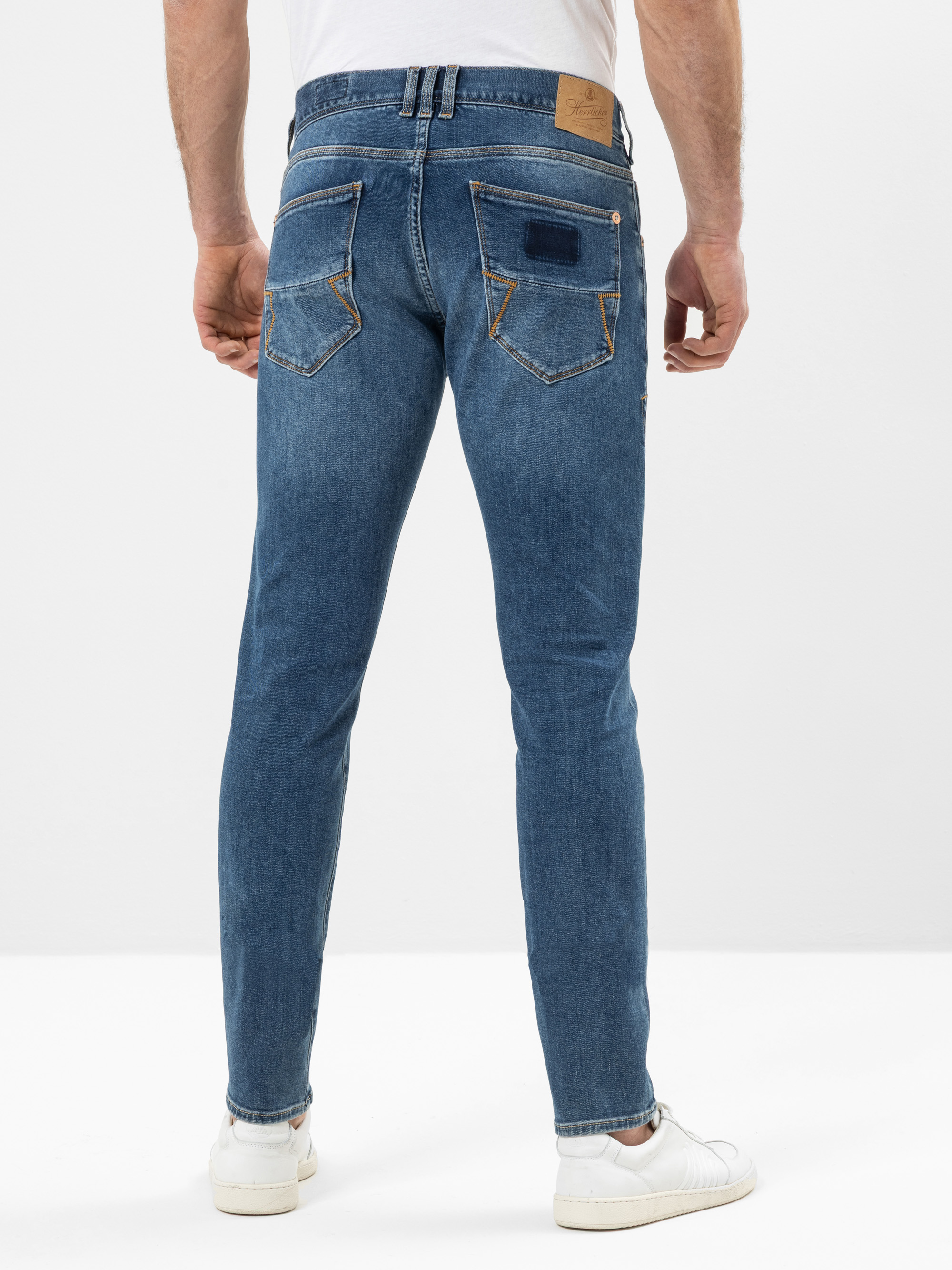 Eine Person, die eine blaue Slim-Fit-Jeans und weiße Turnschuhe trägt, steht mit dem Rücken zur Kamera vor einem schlichten weißen Hintergrund. Die Jeans hat hellbraune Nähte und ein auffälliges Design der Gesäßtaschen.
