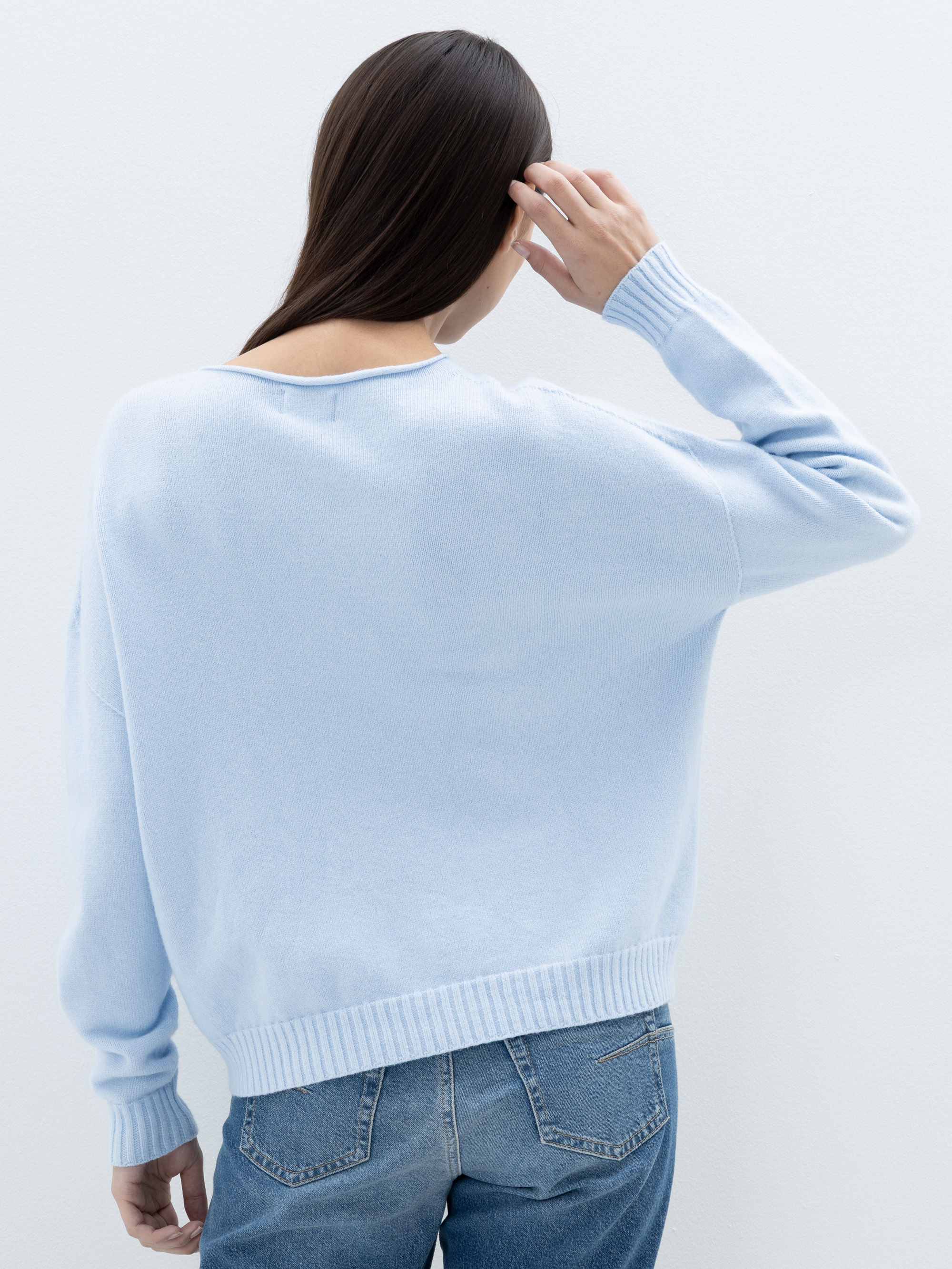 Eine Person mit langem, glattem braunem Haar, die einen hellblauen Pullover und blaue Jeans trägt, wird von hinten gezeigt, wobei eine Hand ihren Kopf vor einem schlichten hellen Hintergrund berührt.