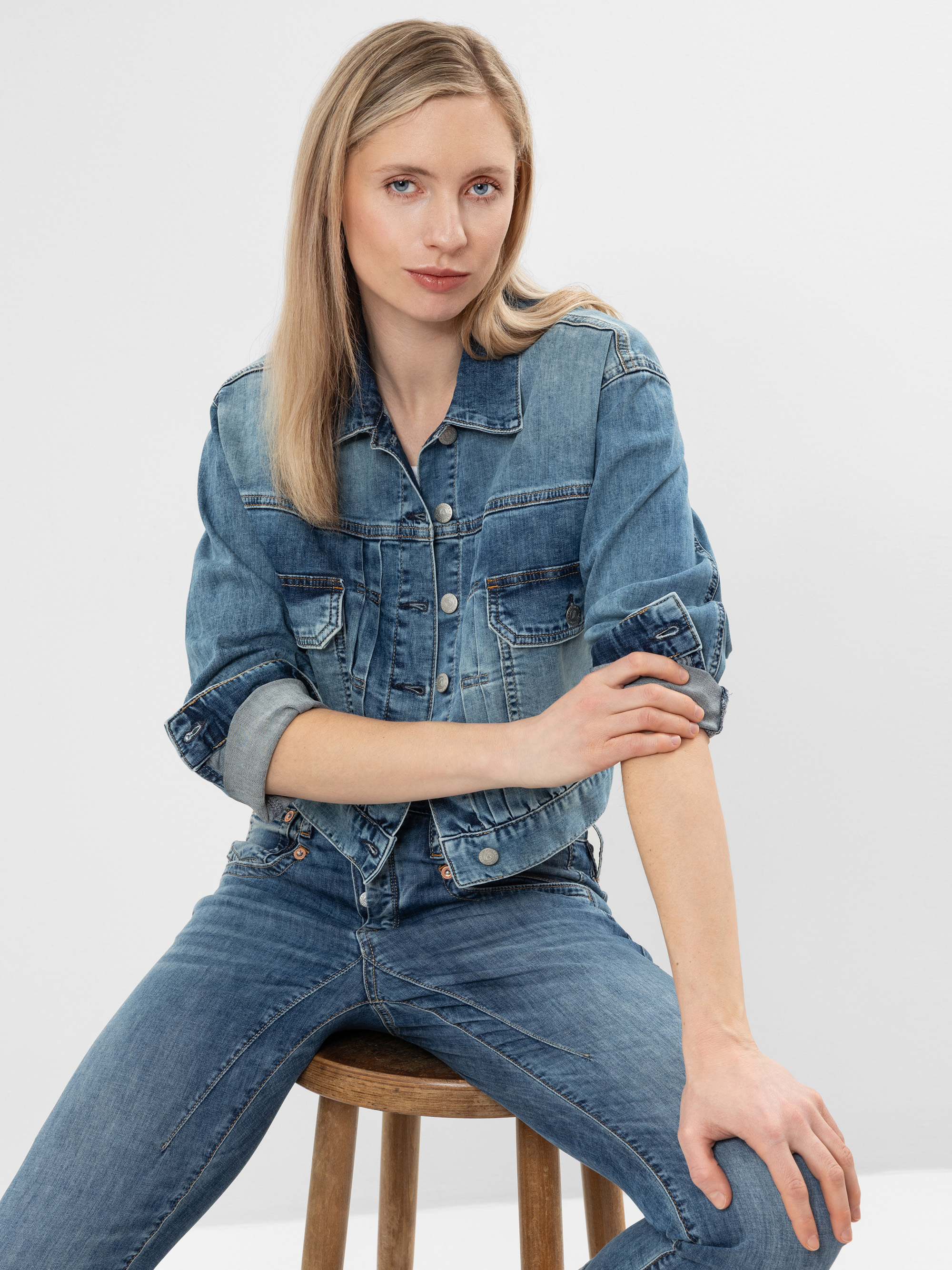 Eine Person mit langen blonden Haaren, Jeansjacke und Jeans, sitzt auf einem Holzhocker vor einem schlichten, hellen Hintergrund und schaut selbstbewusst in die Kamera.