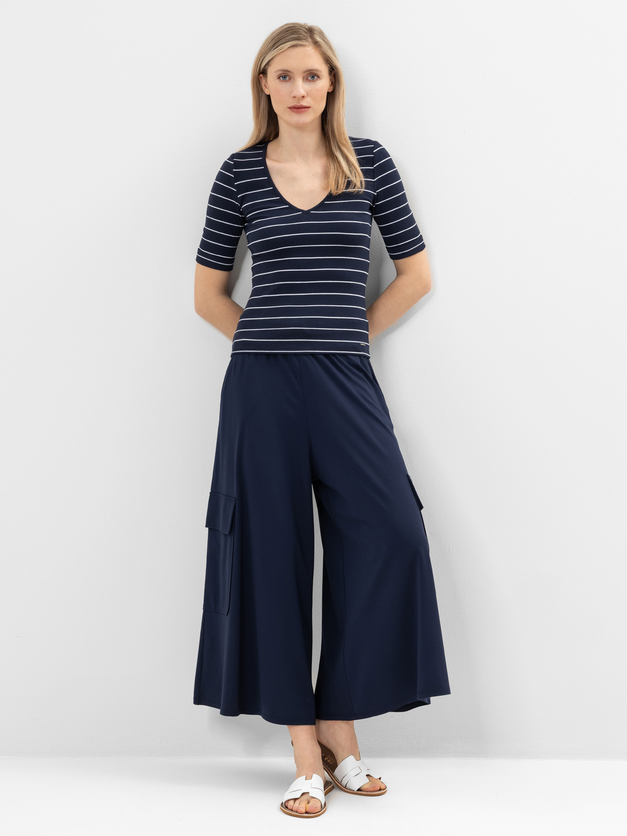 Eine Frau steht vor einer weißen Wand und trägt ein marineblaues Hemd mit V-Ausschnitt und weißen Querstreifen, eine marineblaue Hose mit weitem Bein und Taschen und weiße Slide-Sandalen. Sie hat glattes, hellbraunes Haar und einen neutralen Gesichtsa