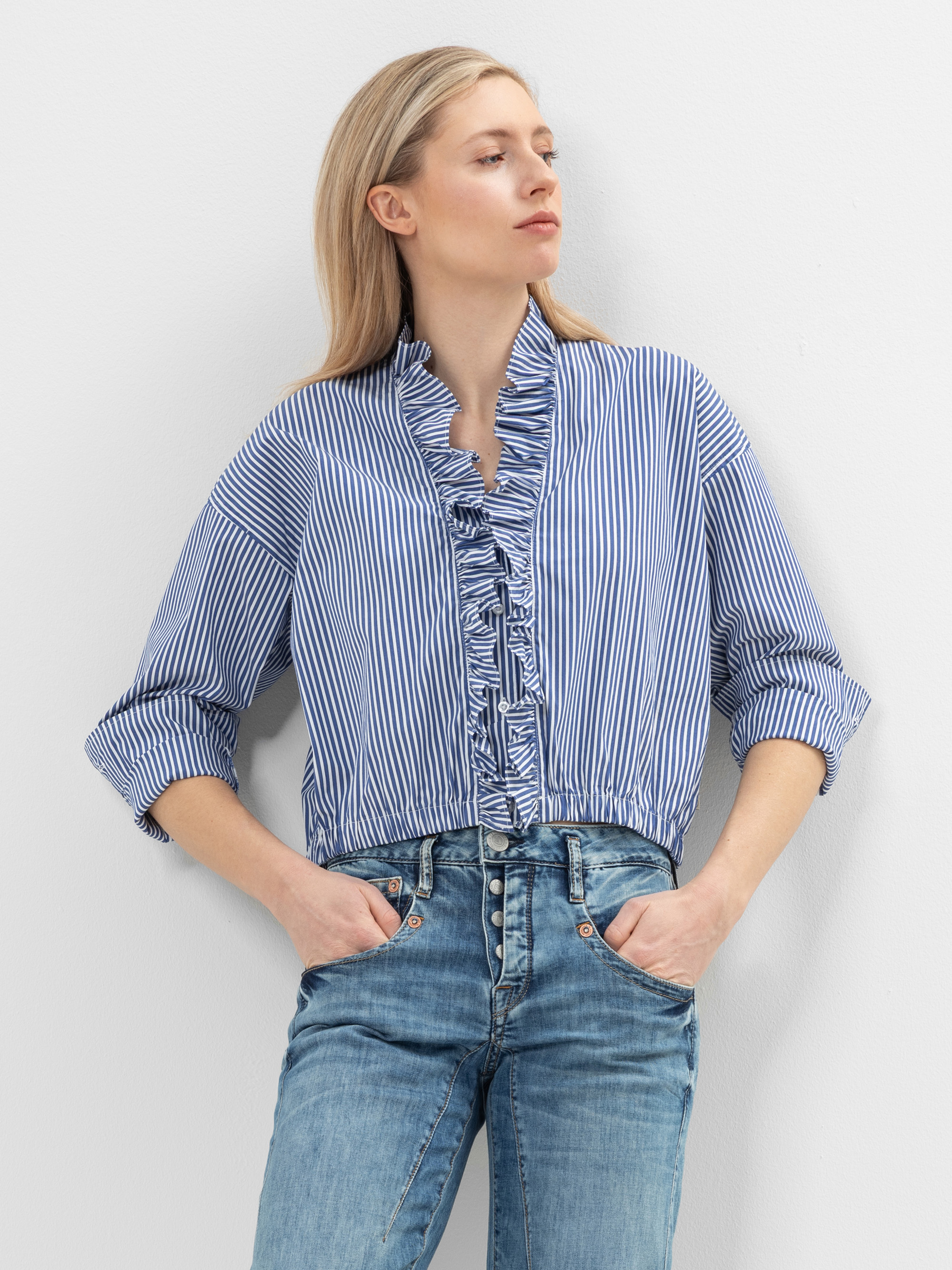 Eine Person steht vor einer weißen Wand und trägt eine blau-weiß gestreifte Bluse mit Rüschen auf der Vorderseite und blaue Jeans. Sie hat helles Haar und die Ärmel sind hochgekrempelt.