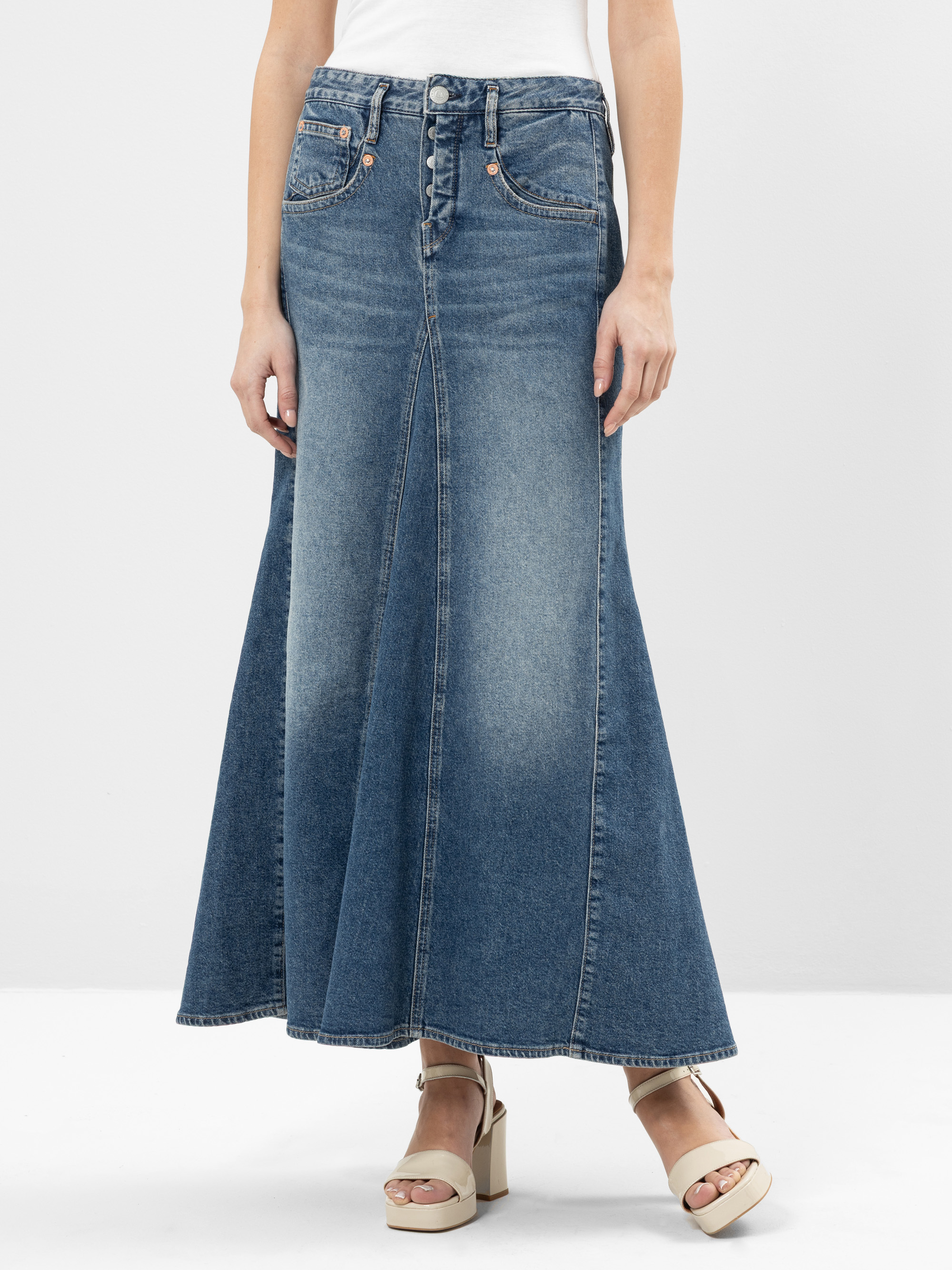 Eine Person, die einen langen, ausgestellten blauen Jeansrock mit Knöpfen an der Vorderseite, ein weißes Oberteil und beigefarbene Plateausandalen trägt, steht vor einem schlichten hellen Hintergrund.