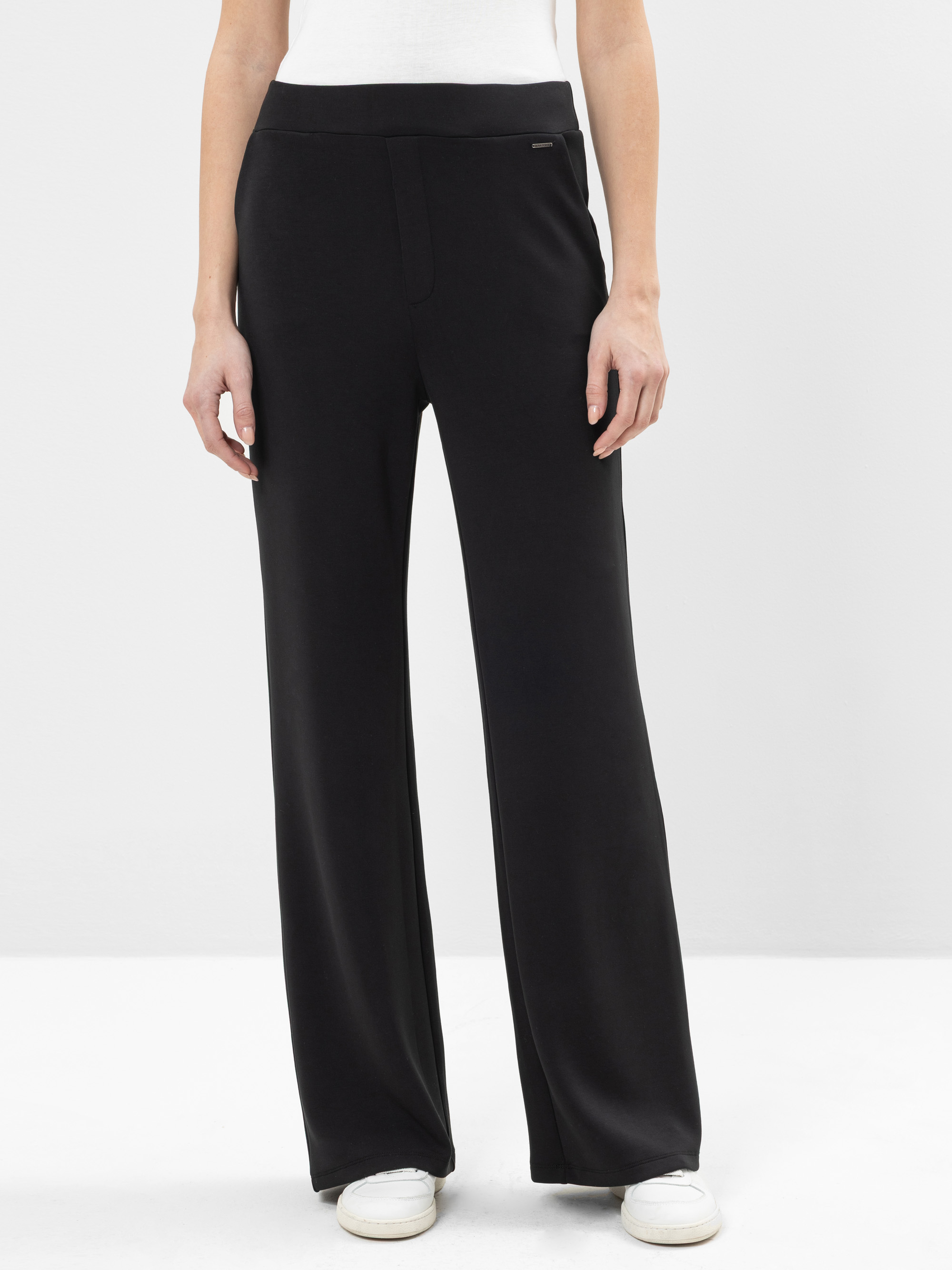 Eine Person, die eine schwarze Hose mit weitem Bein und weiße Turnschuhe trägt, wird von den Schultern abwärts gezeigt und steht vor einem schlichten weißen Hintergrund.