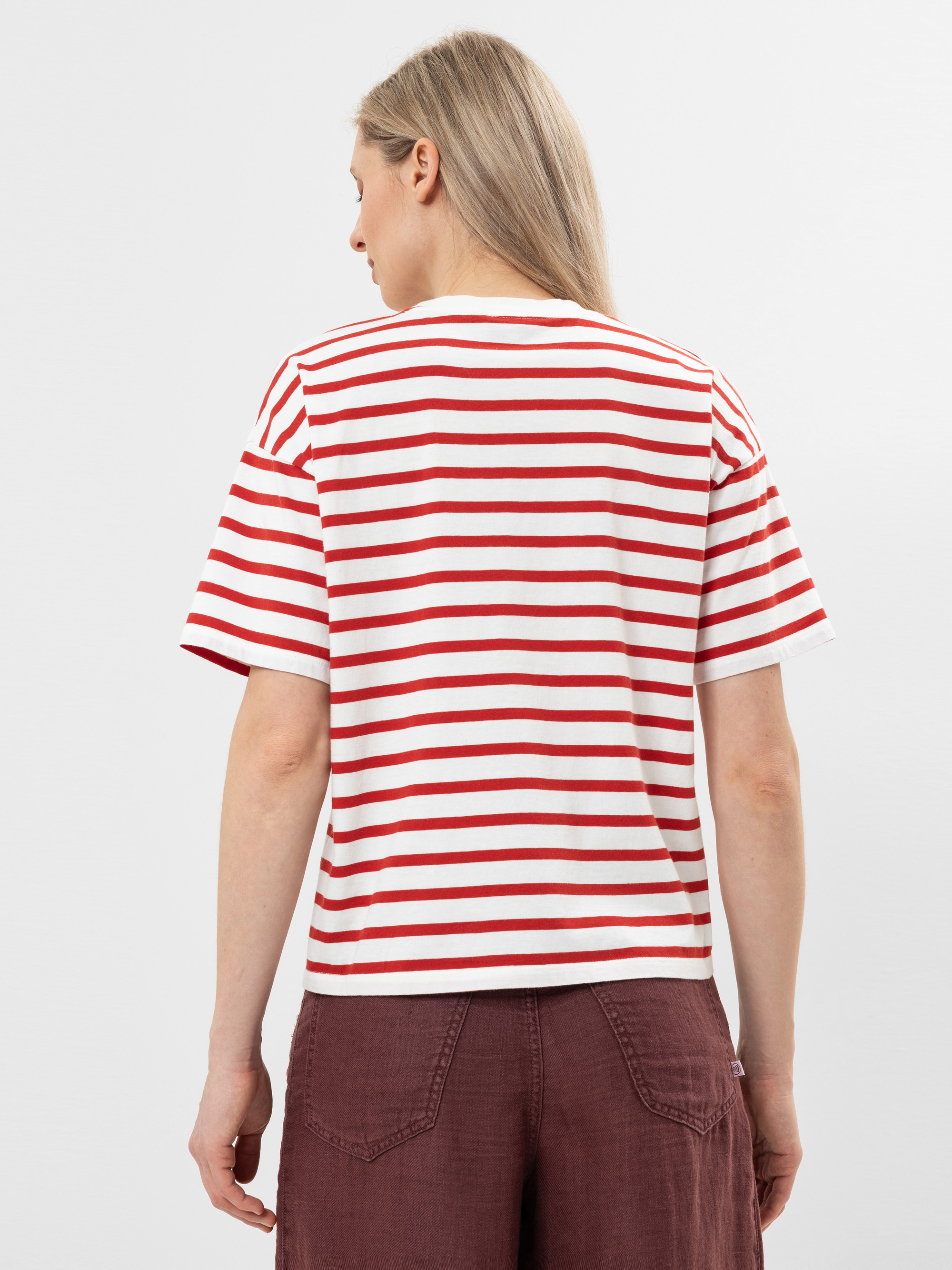 Eine Person mit langen blonden Haaren, die ein rot-weiß gestreiftes kurzärmeliges Hemd und kastanienbraune Shorts trägt, wird von hinten vor einem schlichten hellen Hintergrund gezeigt.