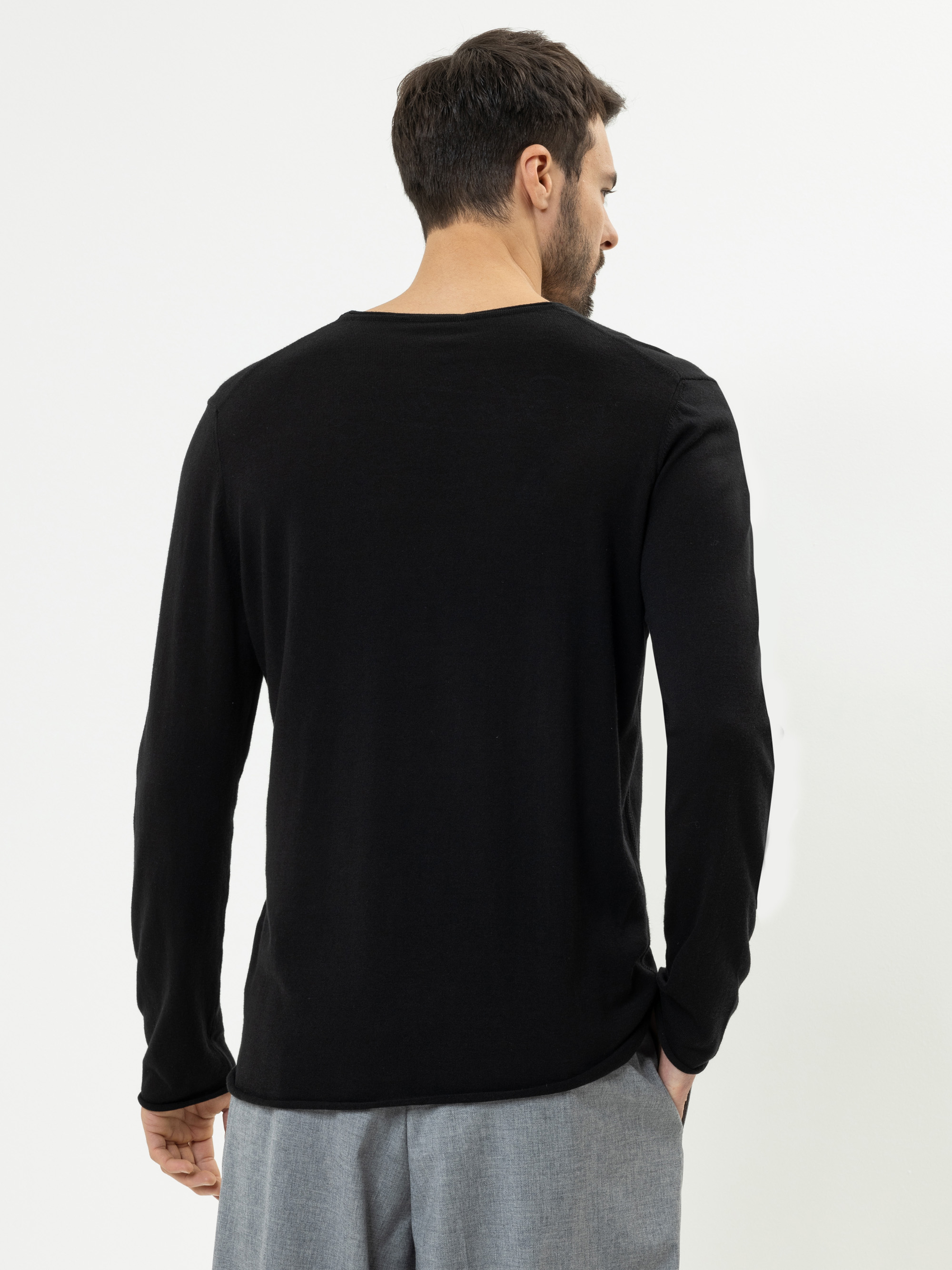 Ein Mann mit kurzen dunklen Haaren blickt von der Kamera weg und trägt ein langärmeliges schwarzes Hemd und eine hellgraue Hose vor einem schlichten weißen Hintergrund.