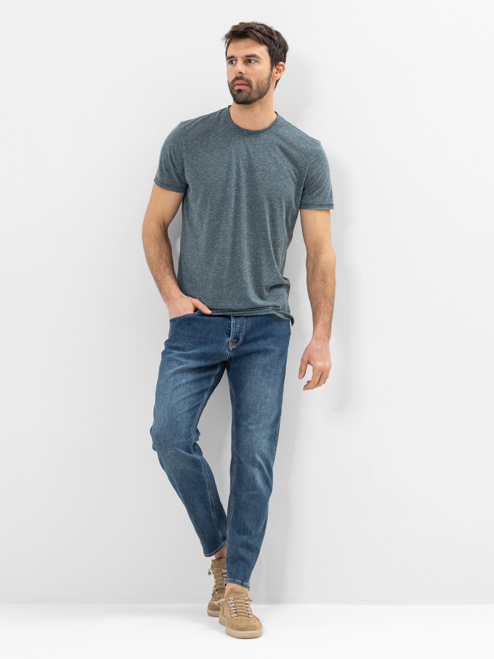 Ein Mann mit kurzen braunen Haaren und einem Bart trägt ein graues T-Shirt, blaue Jeans und beigefarbene Turnschuhe. Er steht vor einem schlichten weißen Hintergrund, hat eine Hand in der Tasche und schaut leicht zur Seite.
