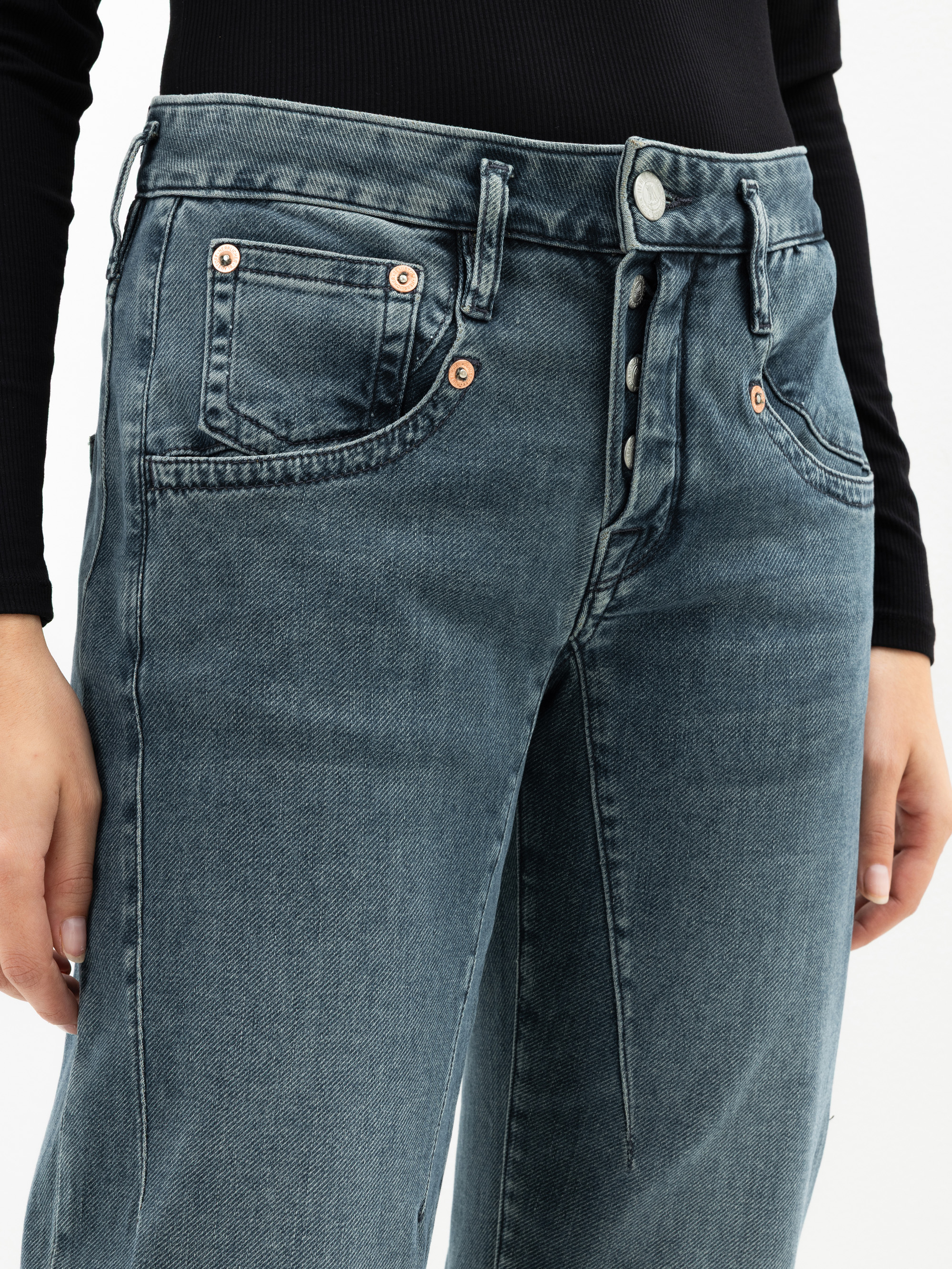 Nahaufnahme einer Person, die eine hochtaillierte blaue Jeans mit freiliegenden Knöpfen und Kupfernieten und ein schwarzes langärmeliges Oberteil trägt. Nur der Oberkörper und die Oberschenkel sind sichtbar.