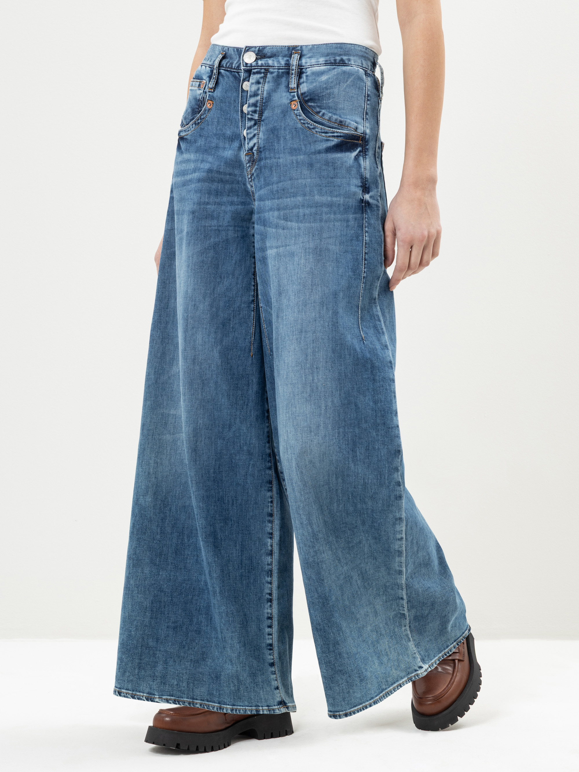Eine Person, die eine blaue Jeans mit weitem Bein und braune, klobige Schuhe trägt, steht vor einem schlichten, hellen Hintergrund. Die Jeans hat eine hohe Taille und markante Nähte. Der Oberkörper der Person ist größtenteils außerhalb des Bildaussc