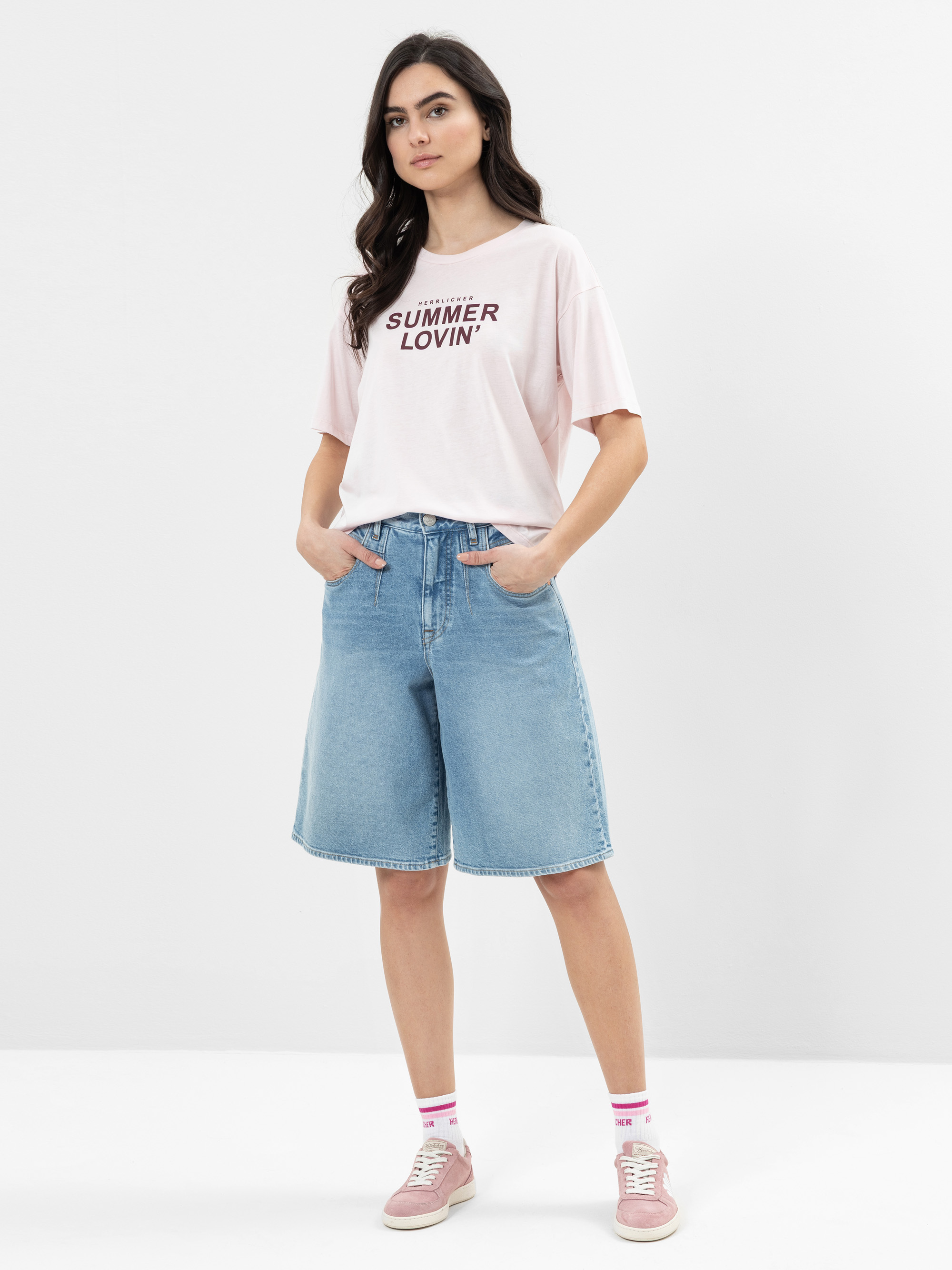 Eine Frau steht mit den Händen in den Taschen da und trägt ein hellrosa SUMMER LOVIN-T-Shirt, hellblaue Bermuda-Shorts aus Denim, rosa Turnschuhe und weiße Socken mit rosa Streifen vor einem schlichten weißen Hintergrund.