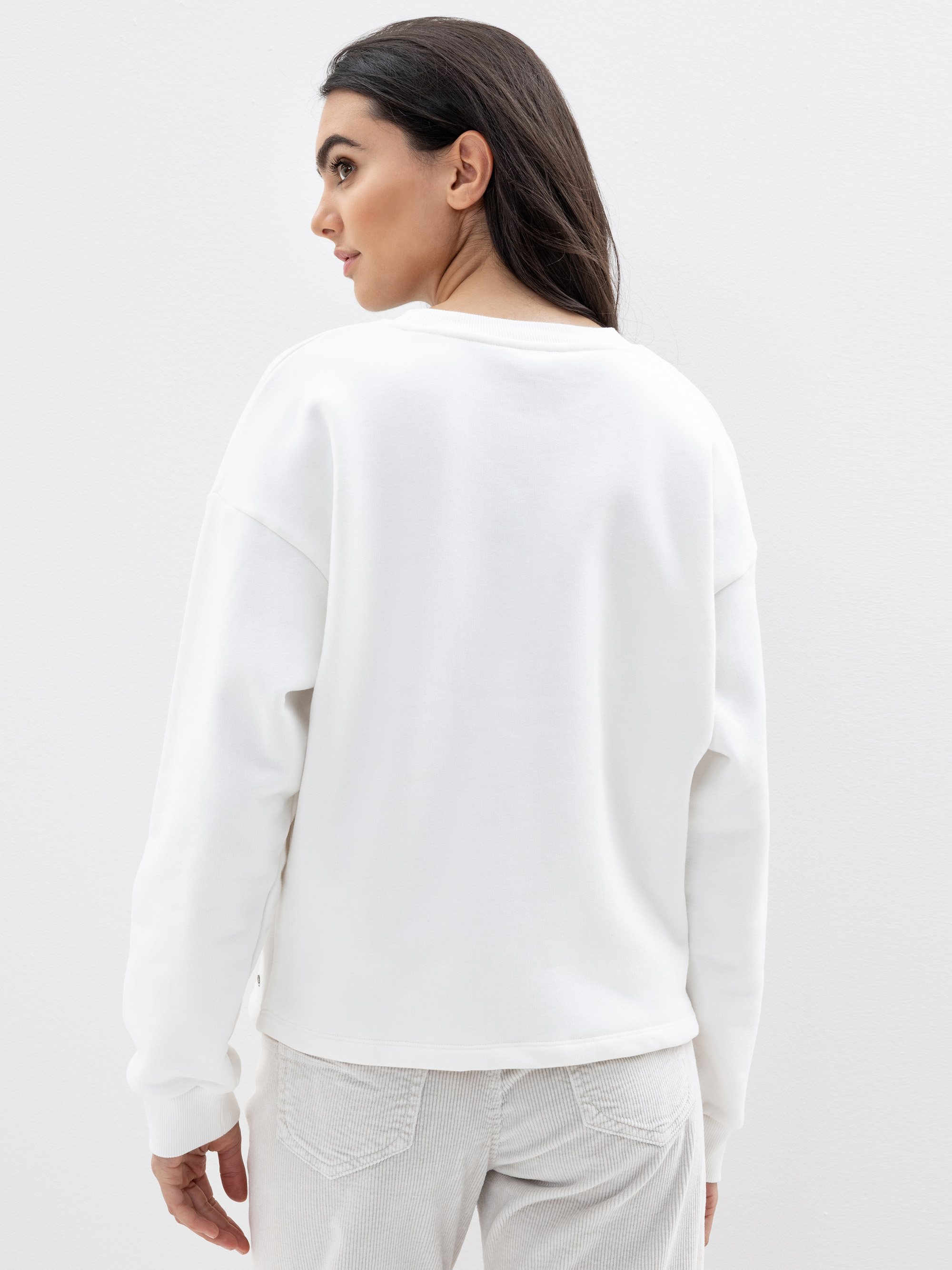 Eine Frau mit langen dunklen Haaren steht, von der Kamera abgewandt, in einem weißen Sweatshirt und einer hellen Hose vor einem schlichten weißen Hintergrund.