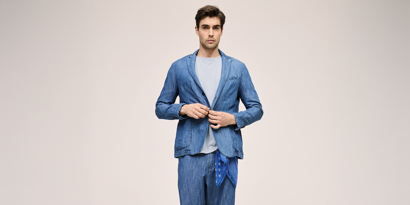Ein Mann steht vor einem schlichten Hintergrund und trägt ein hellblaues T-Shirt, einen blauen Blazer und eine dazu passende blaue Hose. Er knöpft seinen Blazer zu und blickt geradeaus.