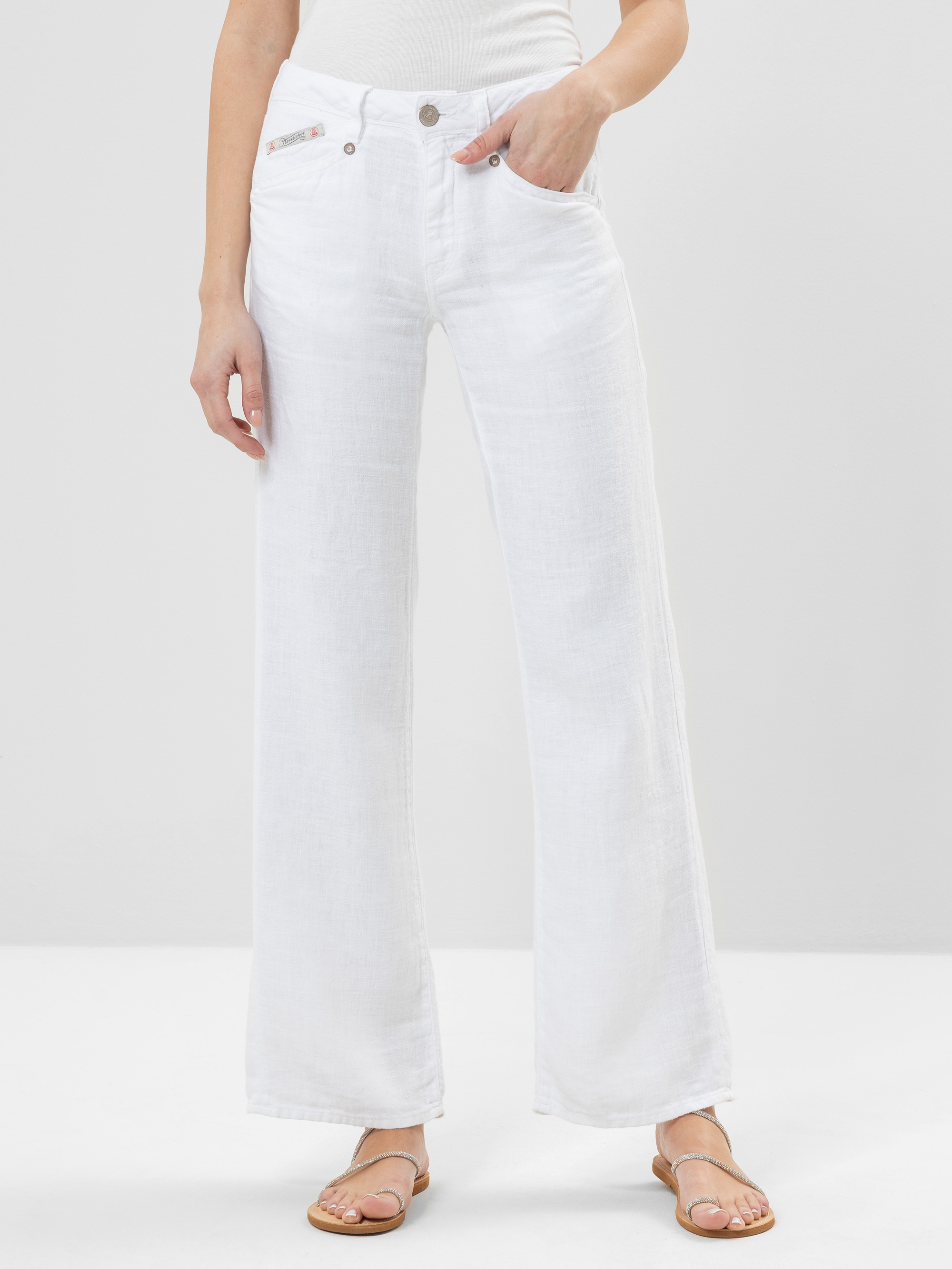 Eine Person, die ein weißes ärmelloses Oberteil, weiße Jeans mit weitem Bein und Sandalen trägt, steht mit den Händen in den Taschen vor einem schlichten hellen Hintergrund.