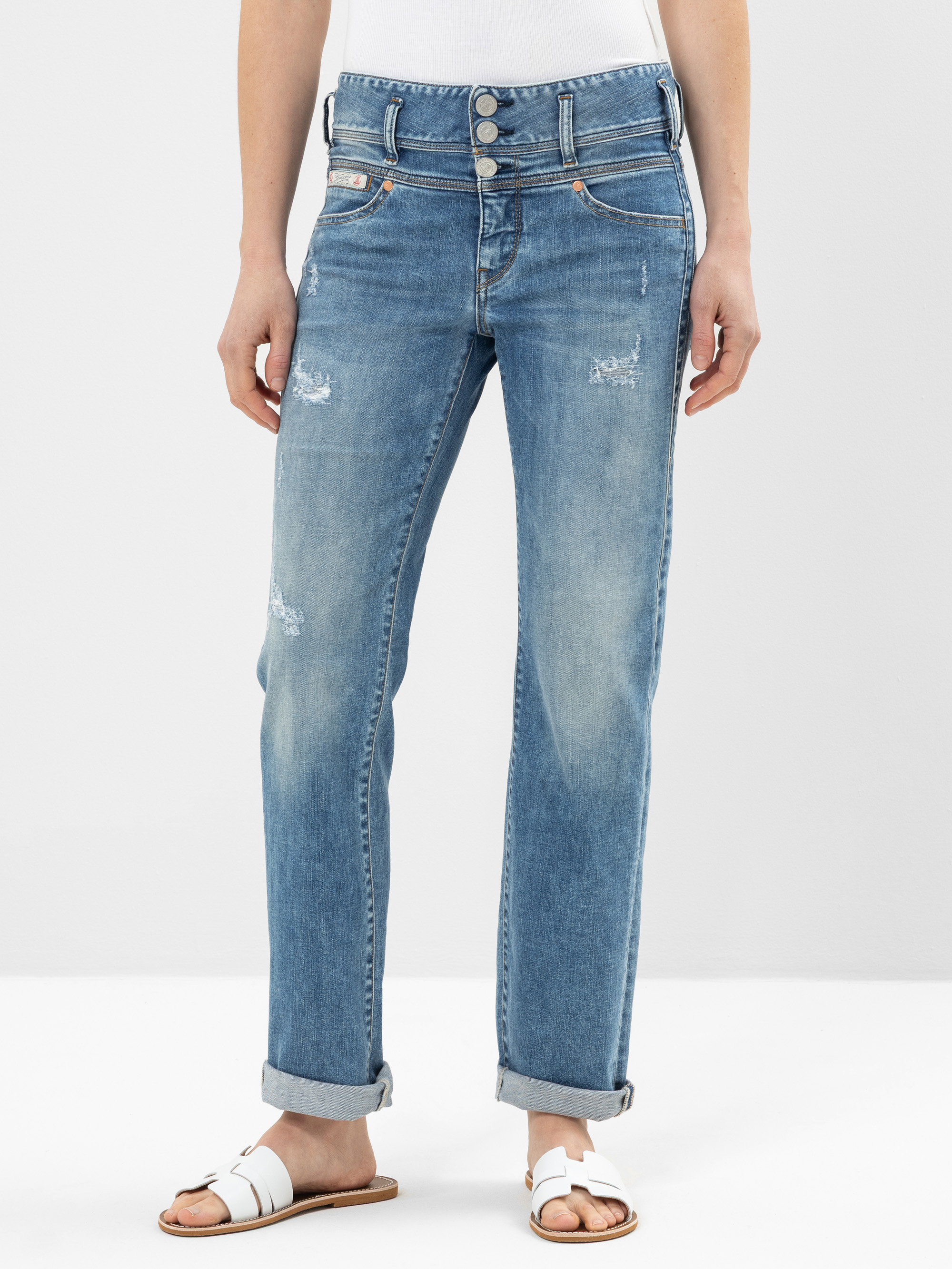 Eine Person, die hellblaue, leicht abgenutzte Jeans mit aufgerollten Manschetten, ein weißes Oberteil und weiße Sandalen trägt, steht vor einem schlichten hellen Hintergrund.