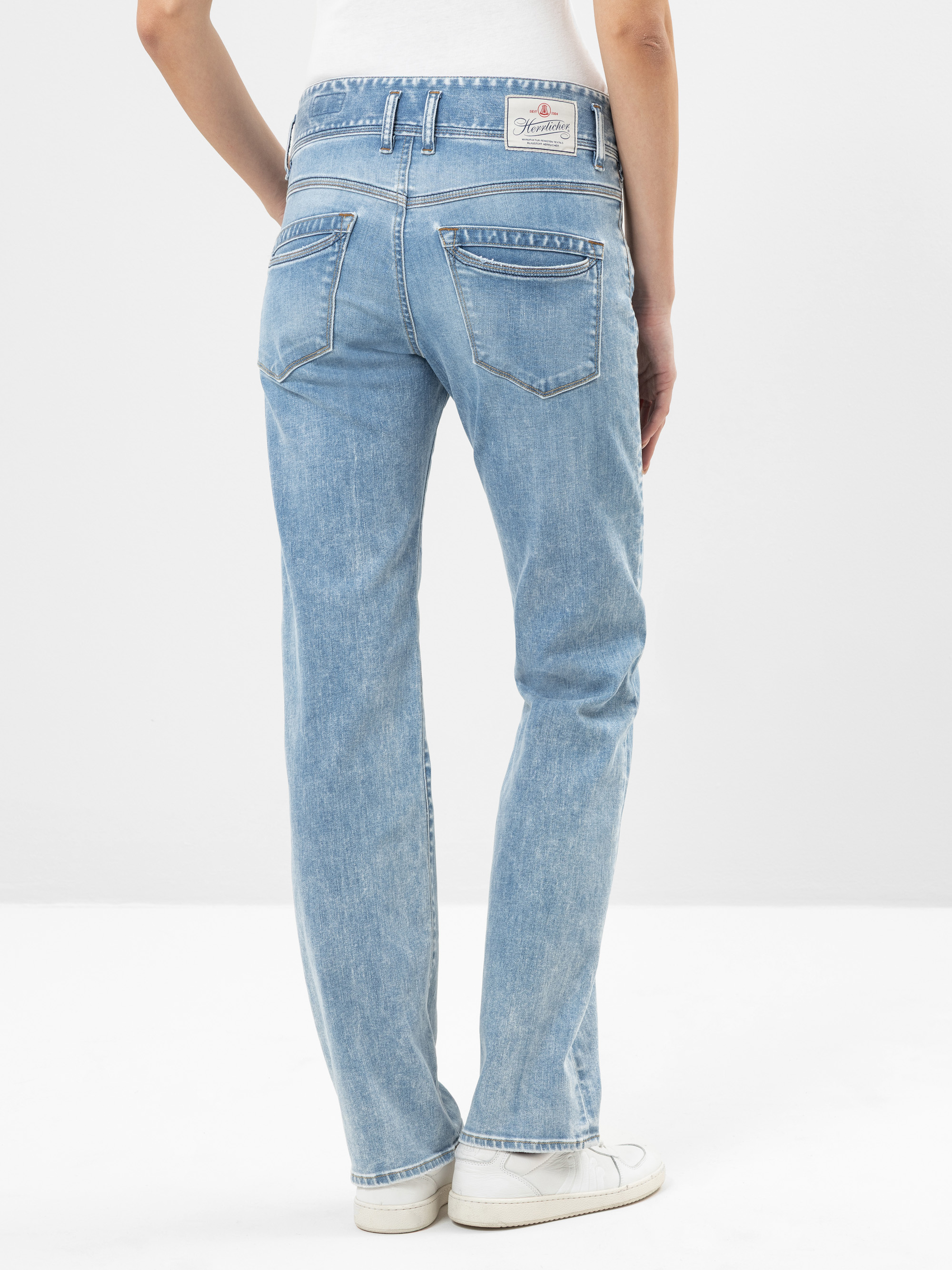 Eine Person in hellblauen Jeans mit geradem Bein und weißen Turnschuhen, von hinten gesehen, steht vor einem schlichten weißen Hintergrund. Die Jeans hat Gesäßtaschen und ein aufgesetztes Etikett am Bund.