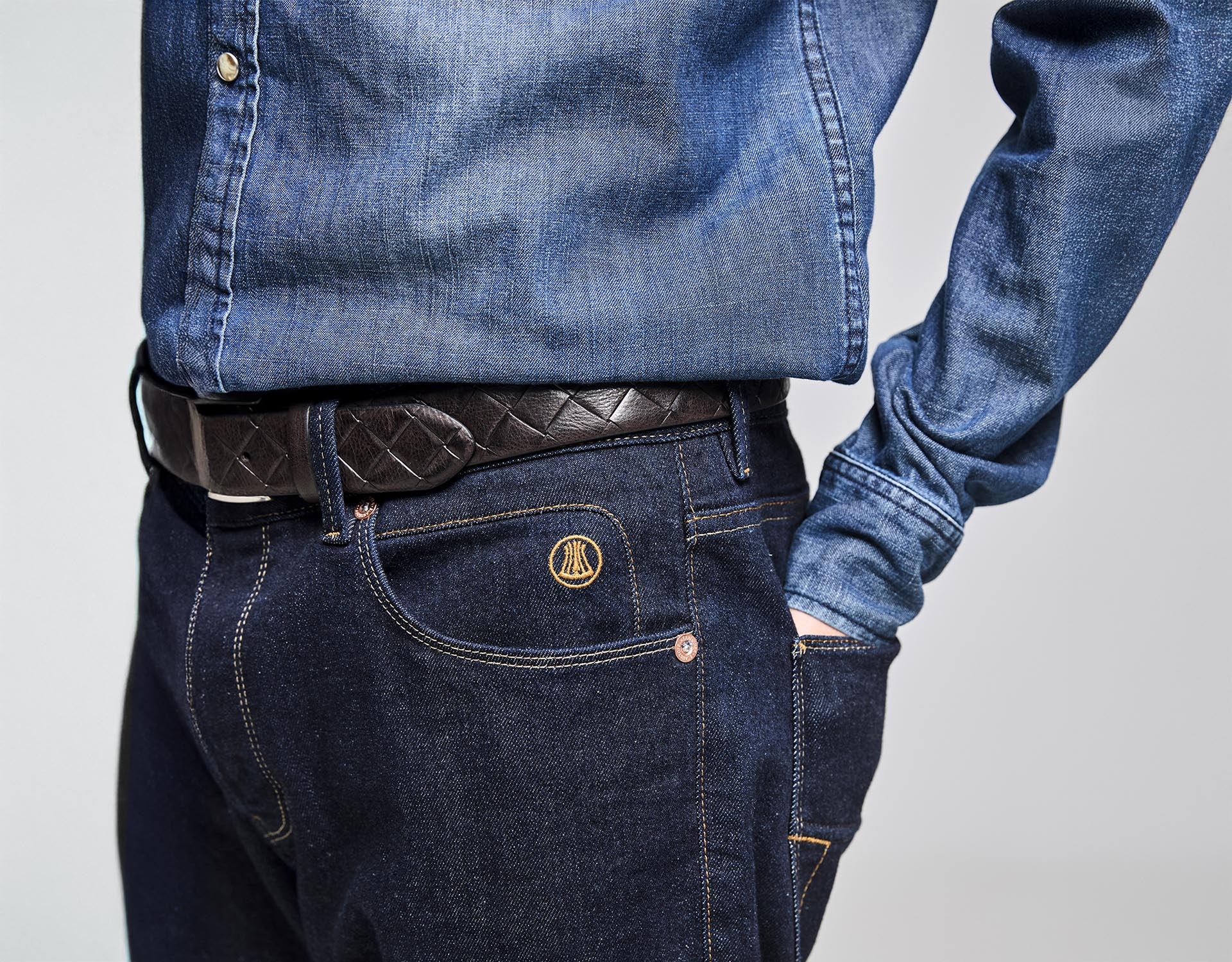 Eine Person, die ein blaues Jeanshemd trägt, das in eine dunkelblaue Jeans mit gelben Nähten gesteckt ist, einen schwarz gemusterten Gürtel trägt und eine Hand in der Tasche hat, wird von den Schultern bis zur Hüfte fotografiert.