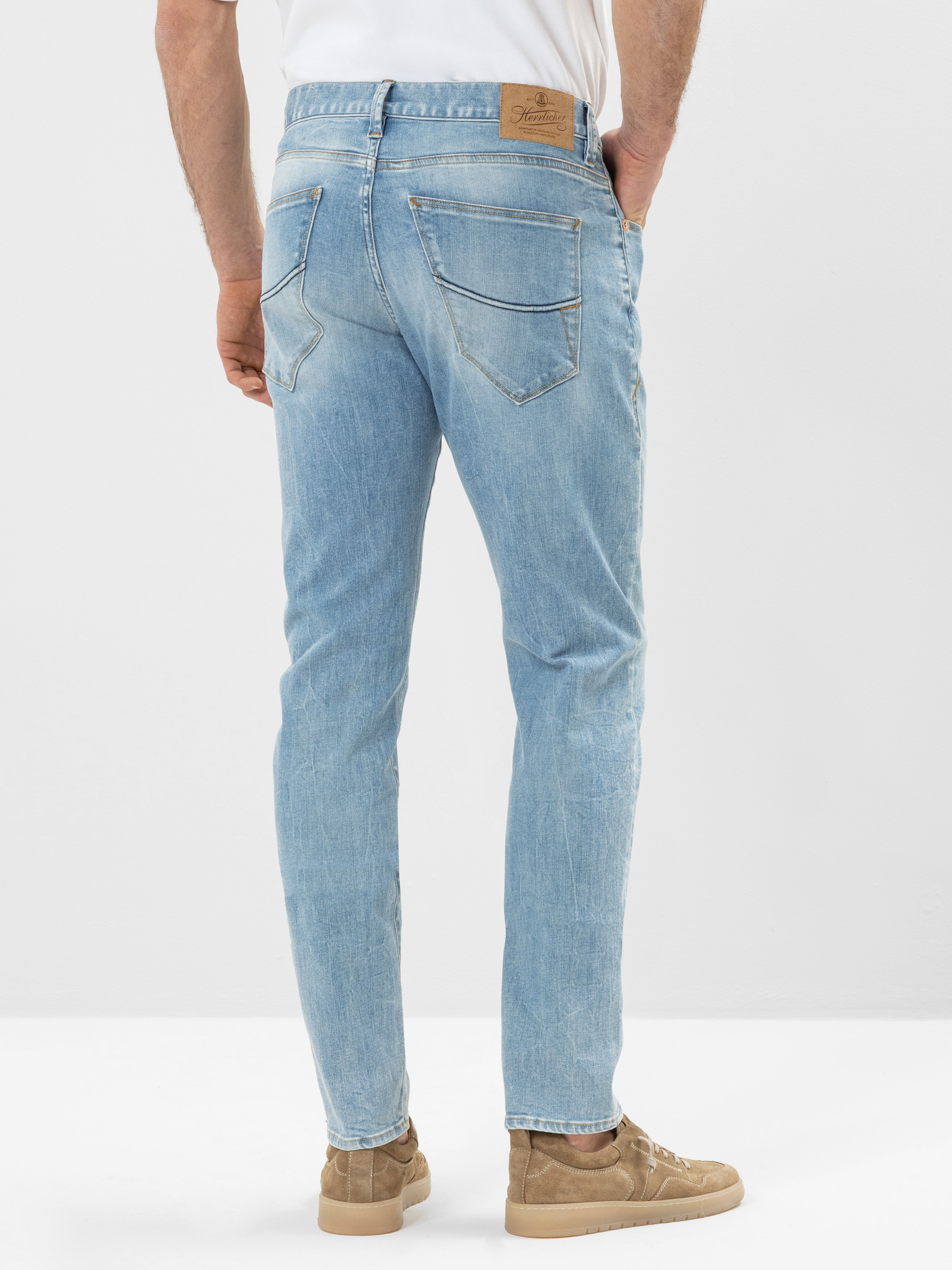 Ein Mann in hellblauen Jeans, weißem Hemd und hellbraunen Turnschuhen steht vor einem schlichten weißen Hintergrund, von hinten gesehen. Seine Hände stecken in den Taschen.