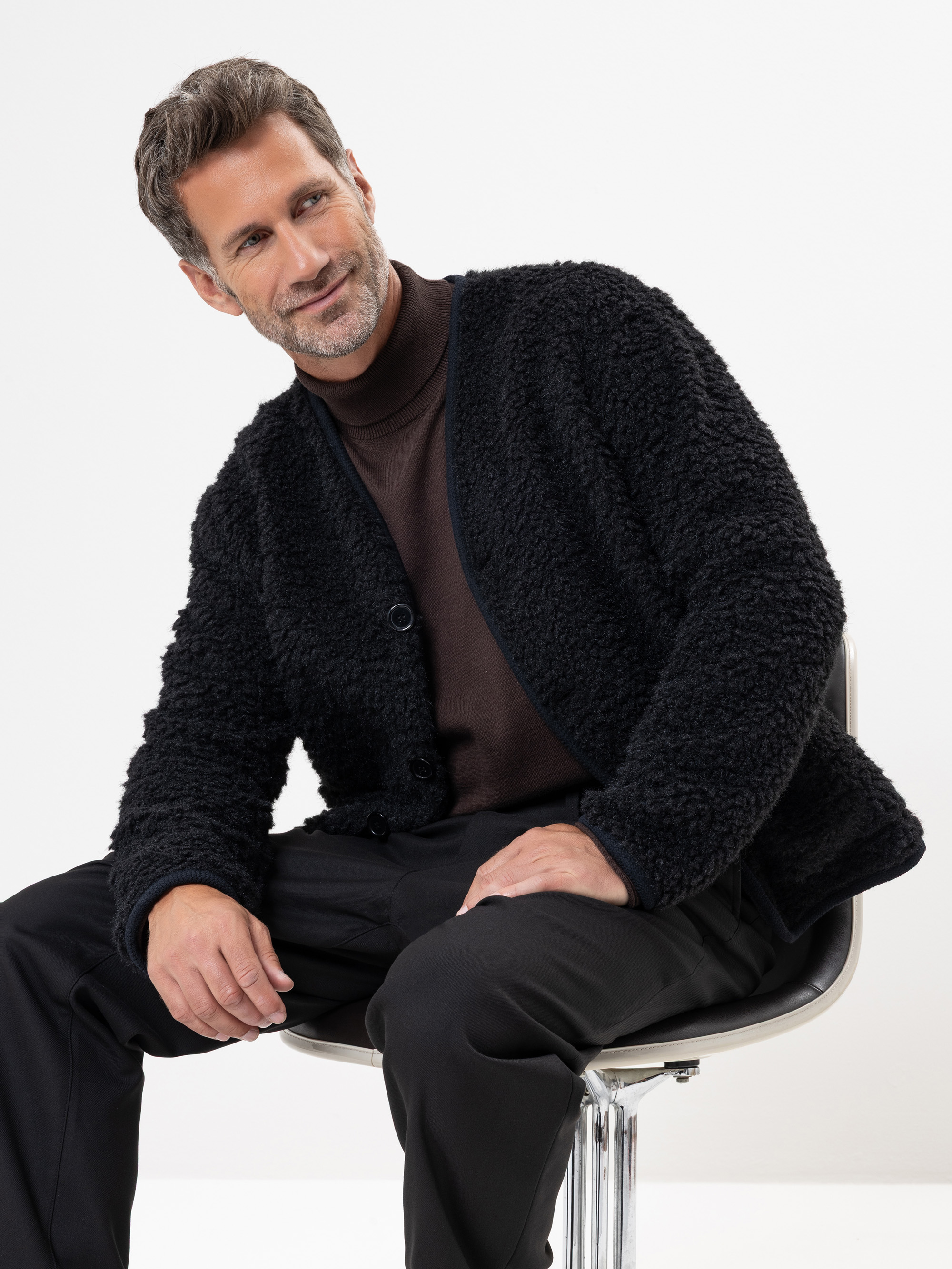 Ein Mann mit kurzen, gewellten braunen Haaren und einem Bart sitzt auf einem modernen Stuhl und trägt eine schwarze, strukturierte Jacke über einem dunklen Rollkragenpullover und eine schwarze Hose. Er lächelt und blickt zur Seite vor einem weißen Hin