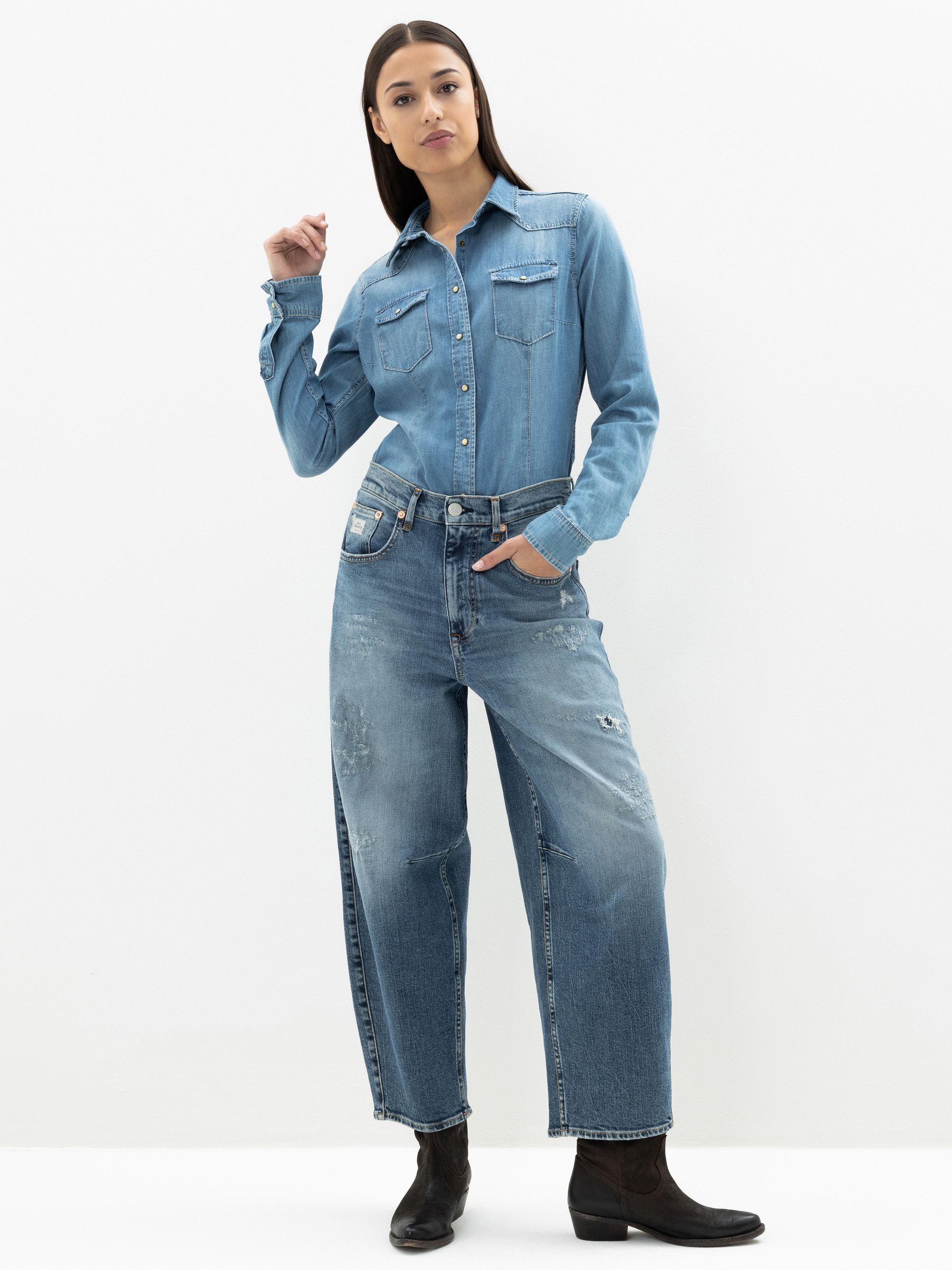Eine Frau steht vor einem weißen Hintergrund, trägt ein hellblaues Jeanshemd und locker sitzende, leicht abgenutzte Bluejeans und hat eine Hand in der Tasche. Sie hat langes dunkles Haar und trägt schwarze Stiefel.