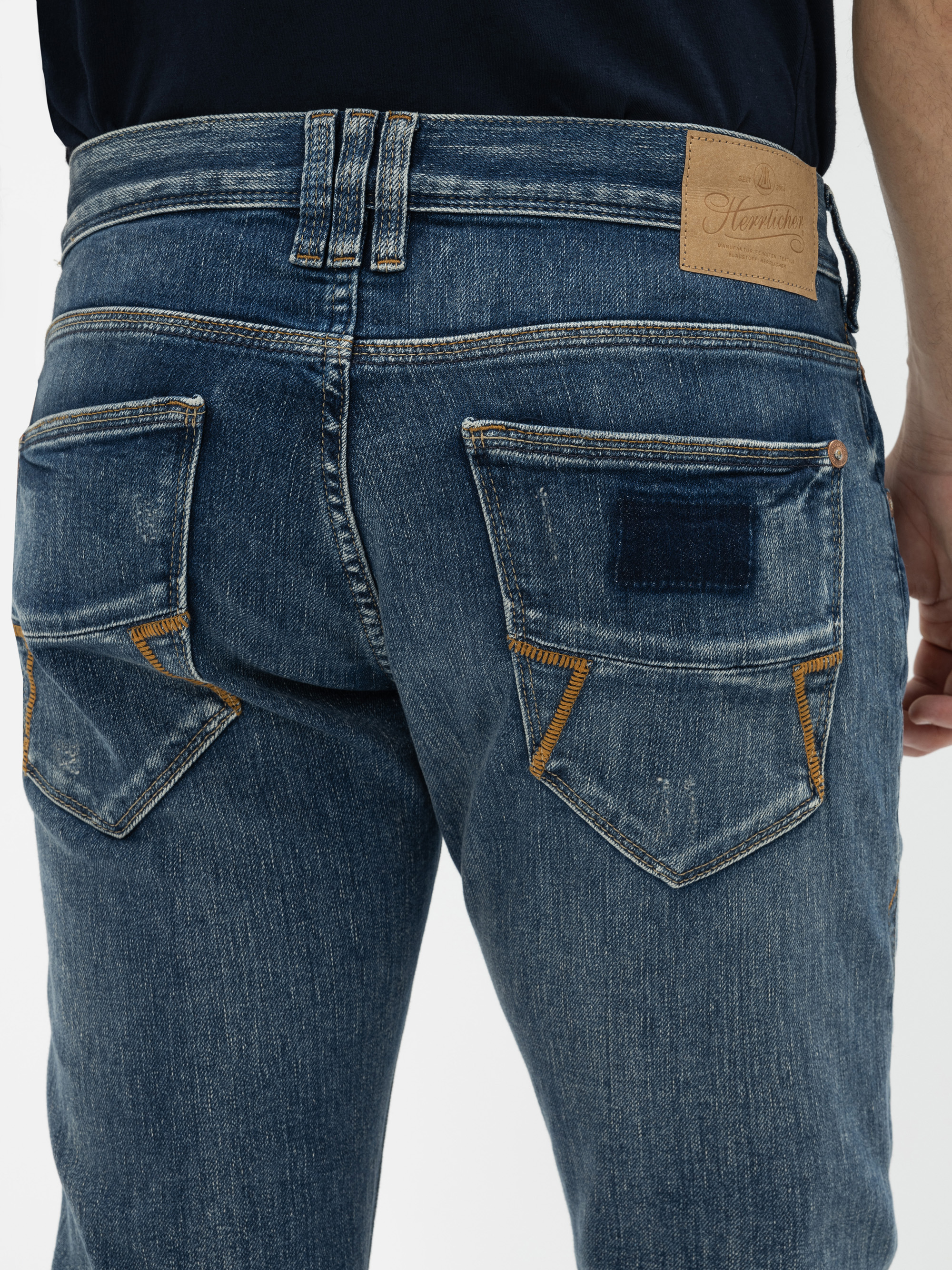 Nahaufnahme des Rückens einer Person, die eine blaue Denim-Jeans mit zwei Gesäßtaschen mit braunen Nähten und einem Markenaufnäher aus Leder am Bund trägt.
